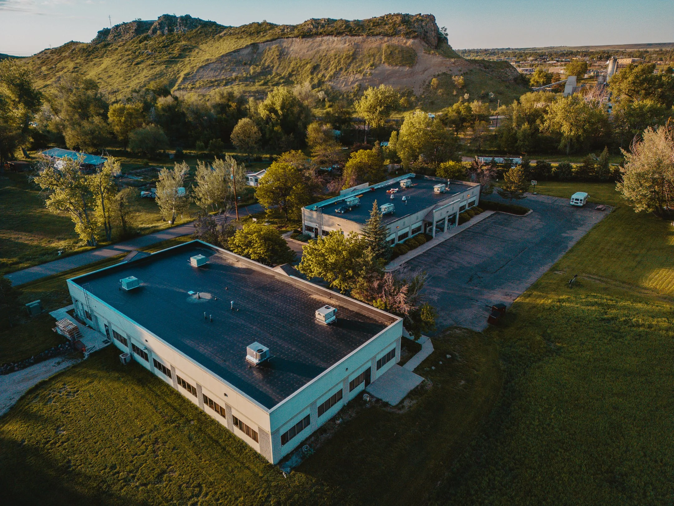 An aerial view of a small campus or office complex with two large, flat-roofed buildings surrounded by greenery, parking lots, and a natural landscape with a hill or small mountain in the background.