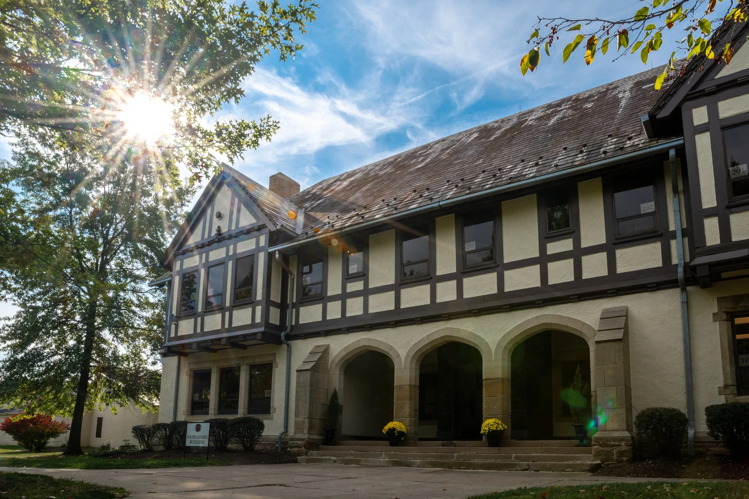 A historic building with Tudor-style architecture, featuring timber framing and stucco walls, with a stone staircase and potted plants at the entrance, surrounded by trees and under a bright blue sky with sunlight.