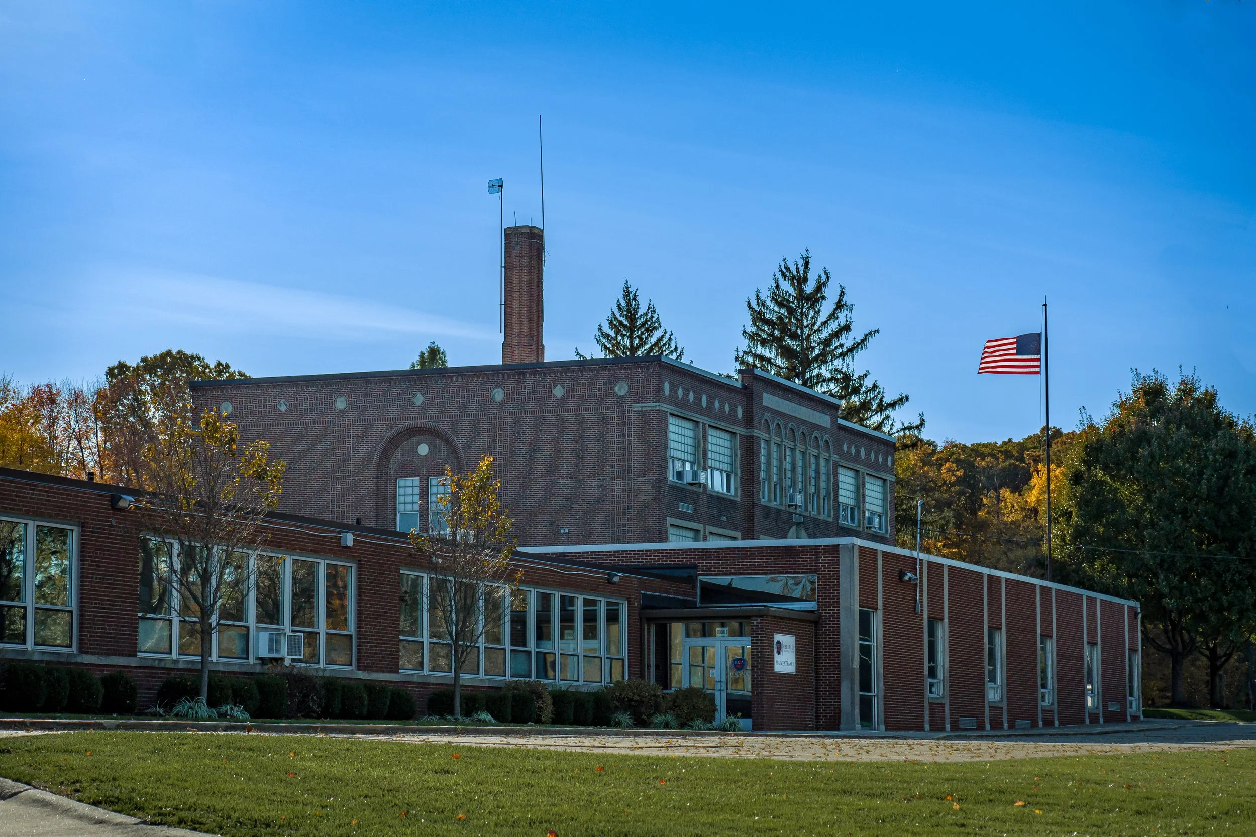 A brick school building with large windows, surrounded by trees with fall foliage, American flags, and a clear blue sky.