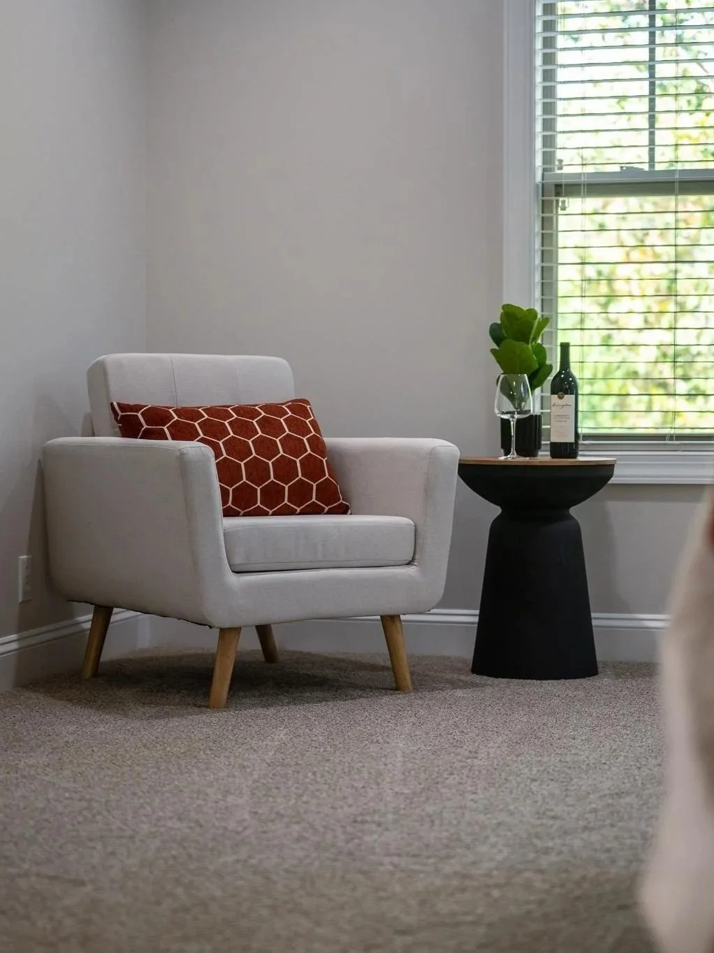 A cozy living room corner with a white armchair, a burnt orange pillow with a hexagon pattern, a black side table with a wine bottle, a glass of wine, and a green plant, next to a window with blinds.