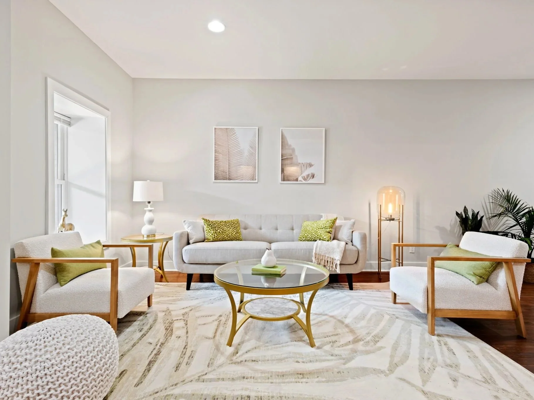A bright living room with white walls and furniture, including a sofa and two armchairs with wooden frames, and green accent pillows. There is a round glass-top coffee table with a gold base in the center, decorated with a white vase and a green book. A white textured ottoman sits on a light-colored patterned area rug. On the left, a window allows natural light in, with a small decorative giraffe on the windowsill. Two framed pictures with abstract designs hang on the wall behind the sofa. To the right, a tall lamp with a clear glass shade and gold stand, along with a potted plant, add warmth to the room.