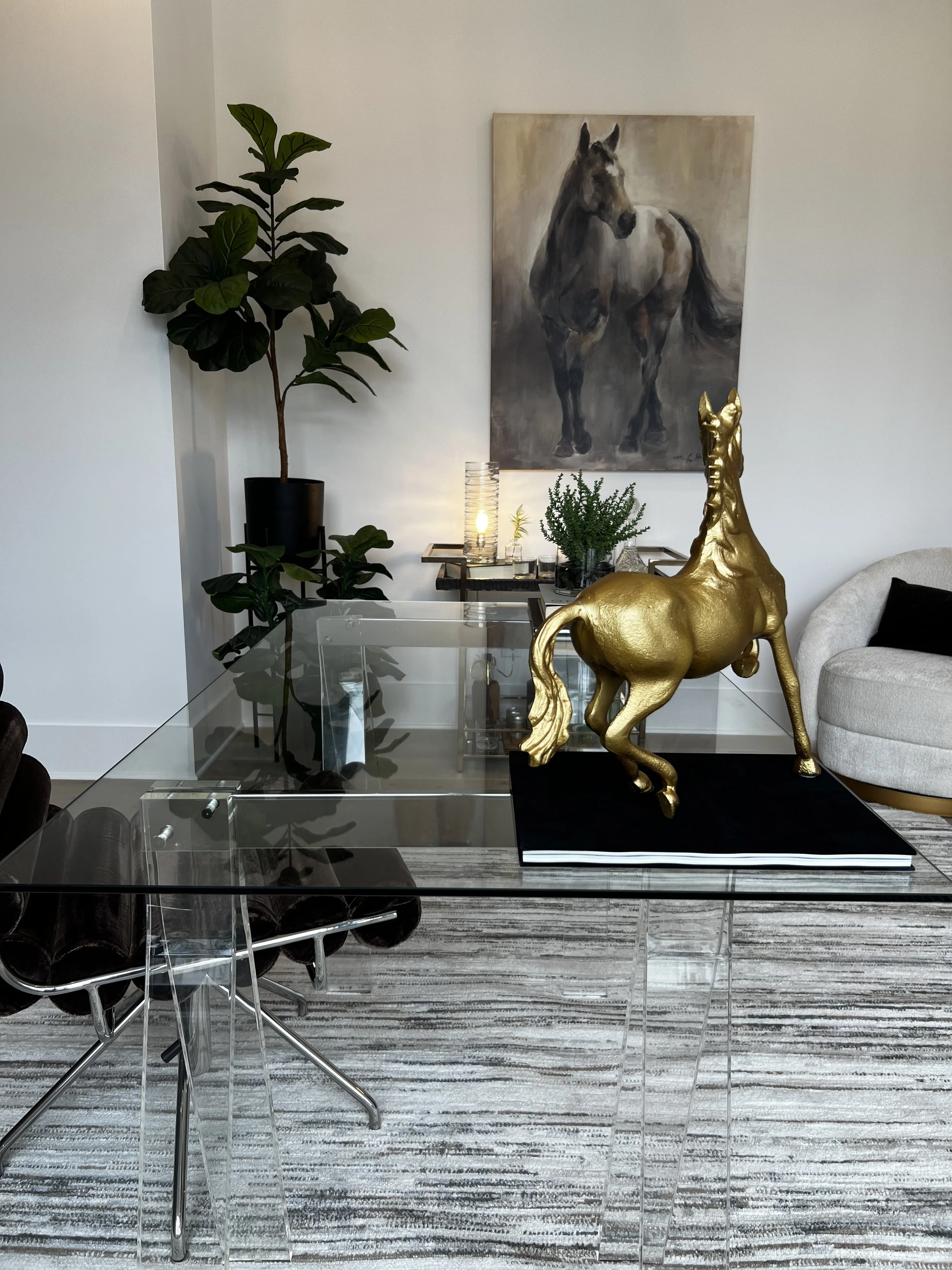 A cozy corner in a room with a blue armchair with an orange patterned pillow, a small wooden side table with a book, a candle, and a lamp, white walls, doorway leading to a balcony with outdoor furniture, and a scenic view of green trees and blue sky outside.