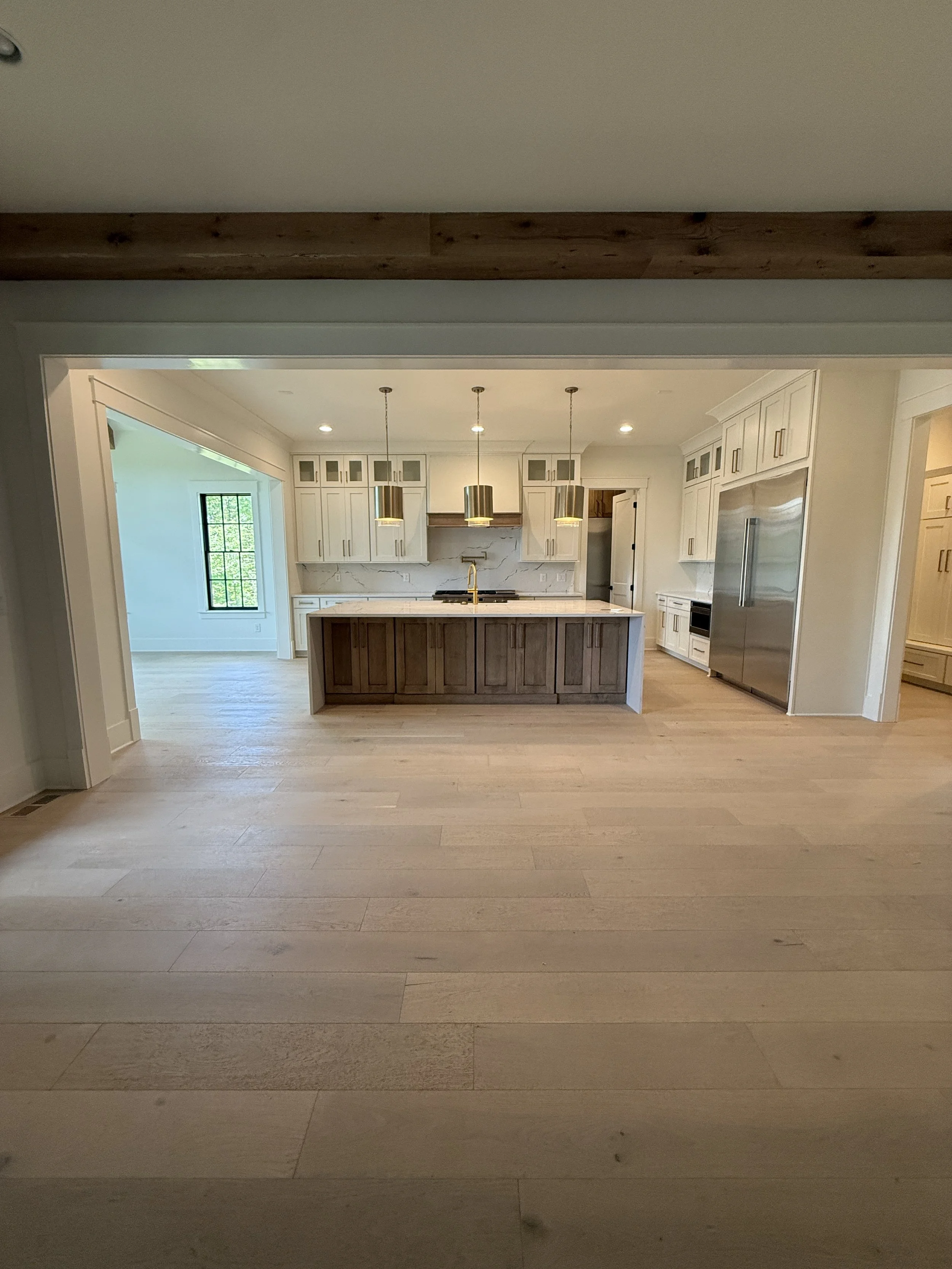 Living room with wooden coffee table, black sculpture, and sofa with green and brown pillows, adjacent to a brick fireplace and a large window, with a kitchen in the background.