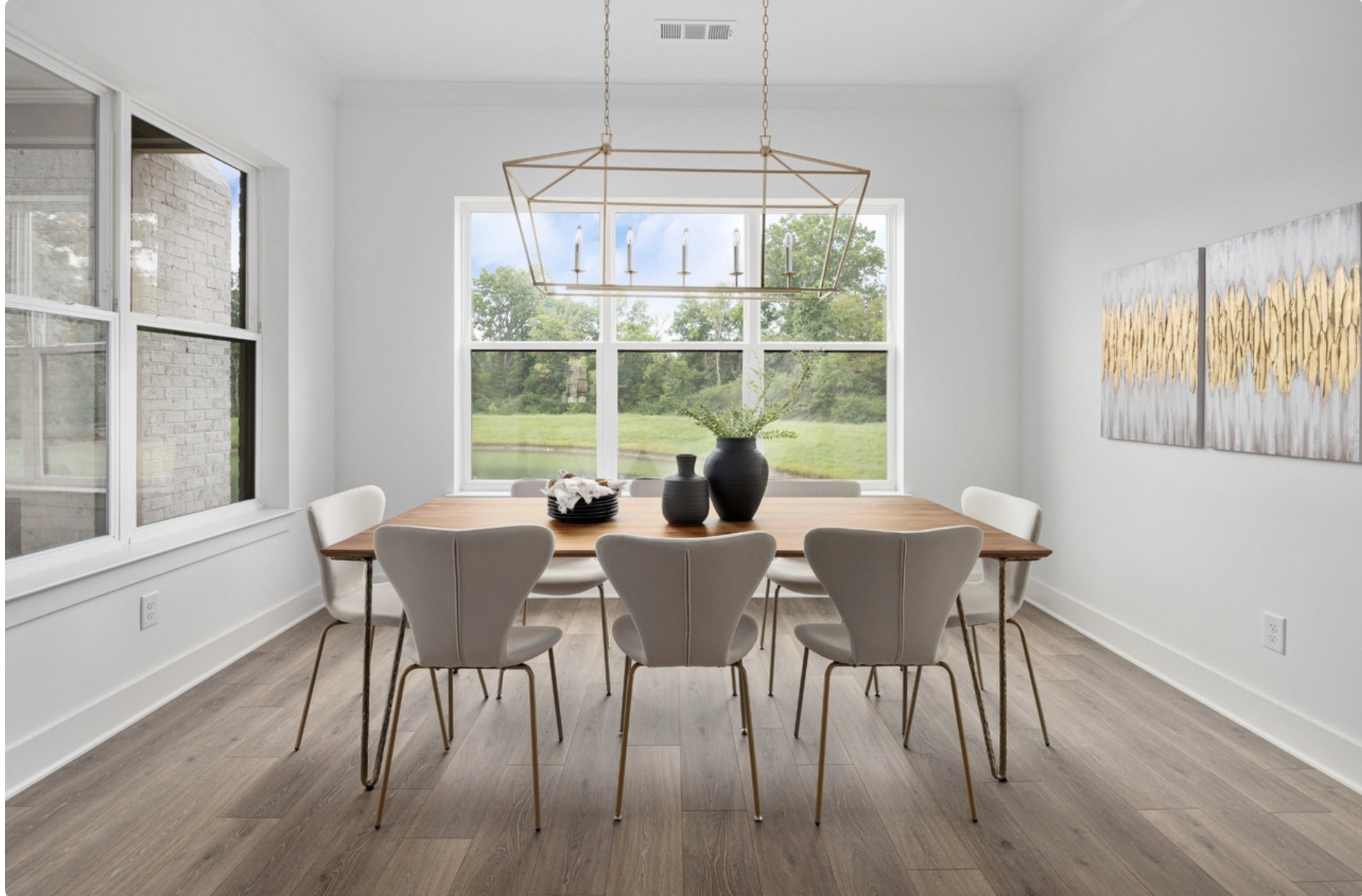 Empty modern dining room with a wooden table, six white chairs, a large window showing a green outdoor landscape, a black vase with greenery, and a geometric gold chandelier.
