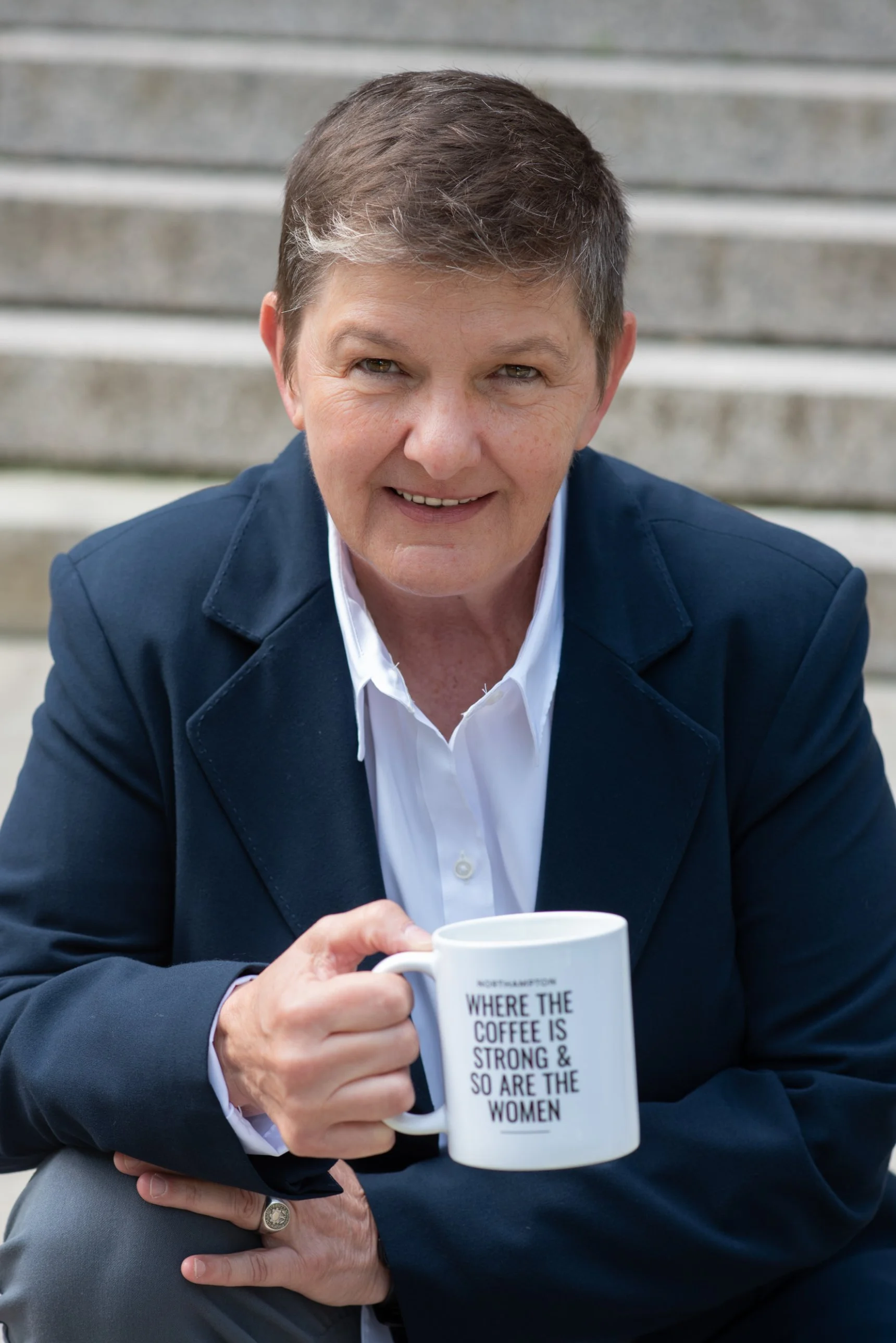 Toni Bator smiling on stone steps, holding a mug that reads Where the coffee is strong and so are the women