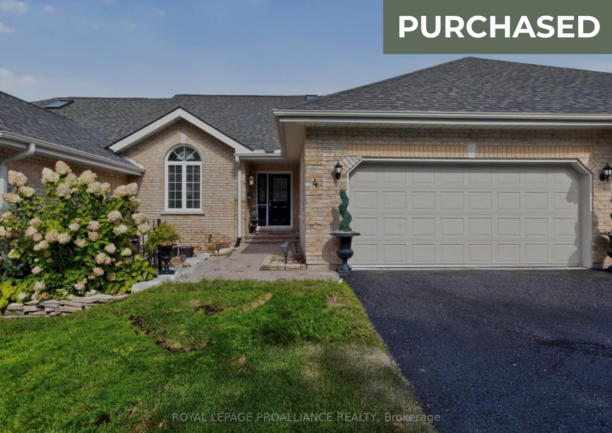 Front view of a brick house with a gray garage door, arched window, and landscaped garden with flowers and green lawn.
