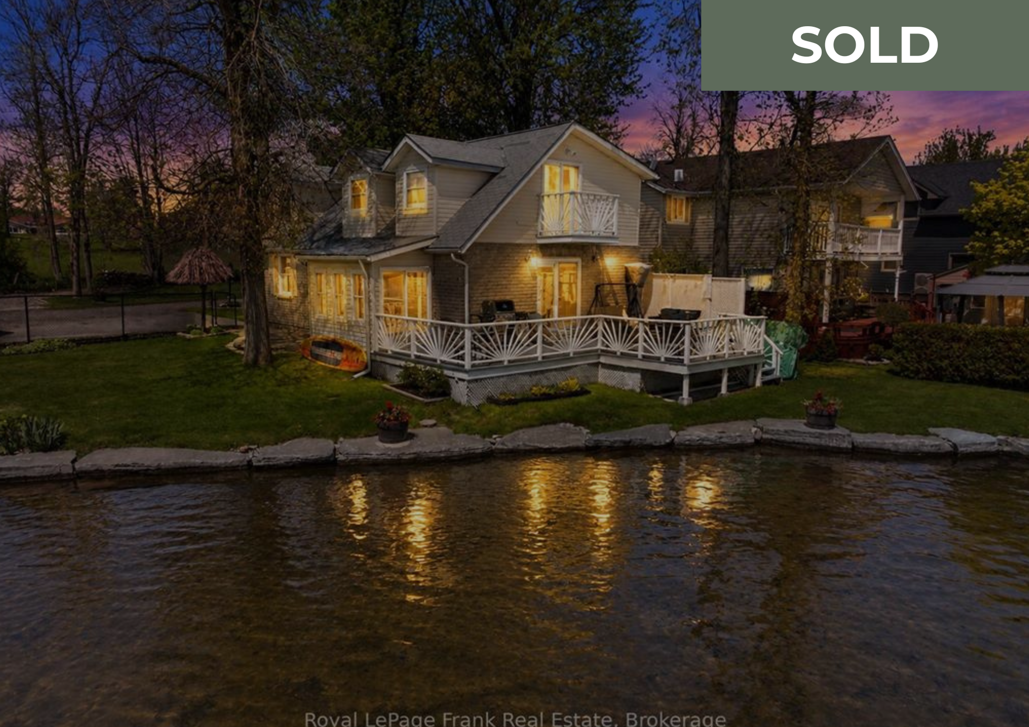 A two-story house with warm yellow interior lights, situated by a river at dusk. The house features a brick lower level and a wooden upper level with a small balcony. Surrounding the house are trees, a grassy yard, and a stone pathway.