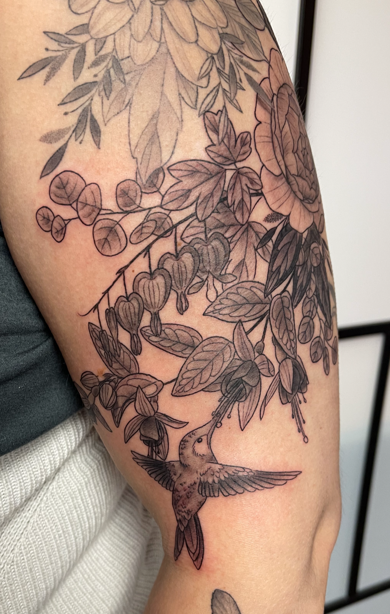 Tattoo of a hummingbird with a flower and leaves on the arm.