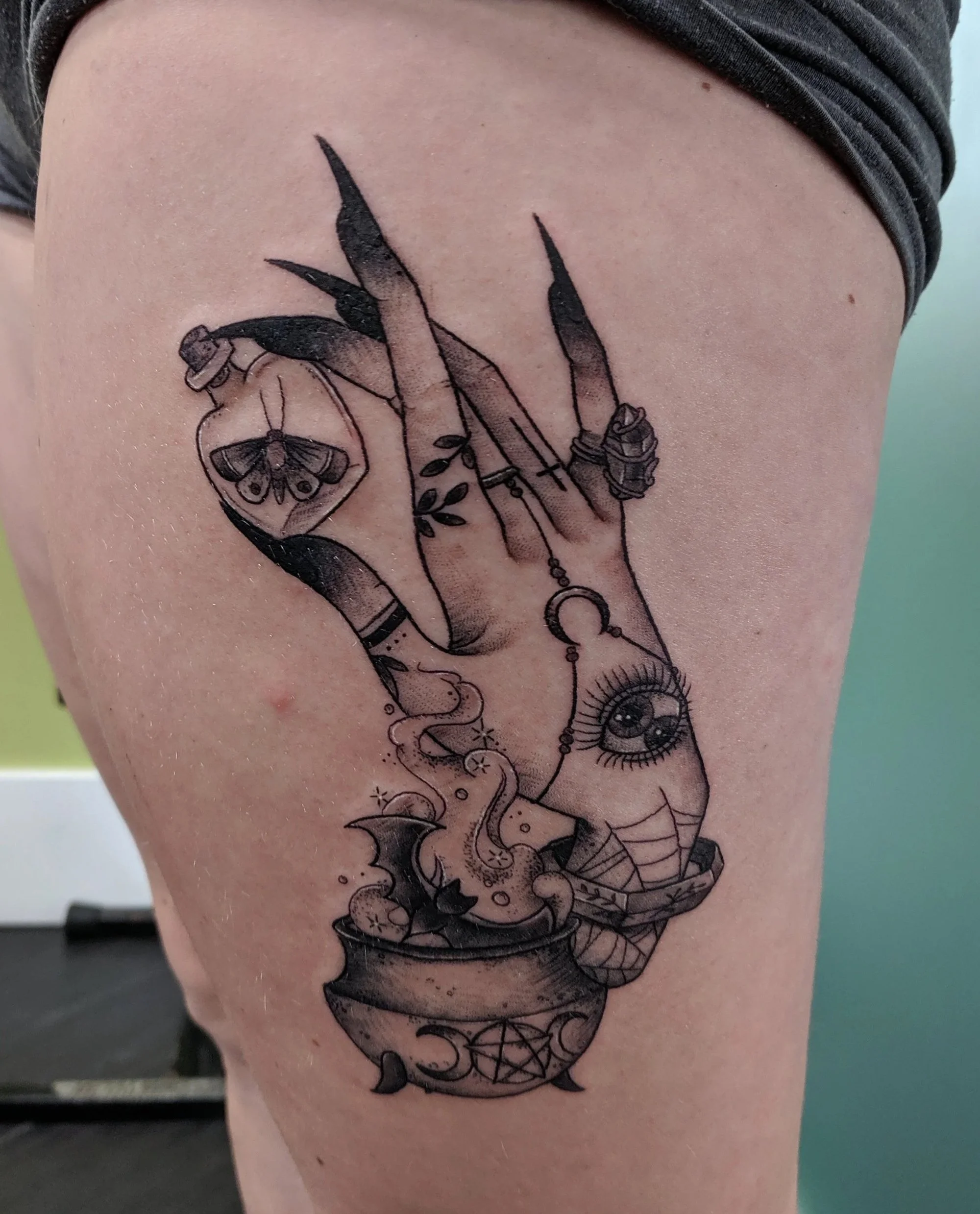 Tattoo of a hand with long black nails holding a vial with a moth inside, an eye on the wrist, and a crystal ball with symbols and a pentagram at the bottom, surrounded by smoke and magical symbols.