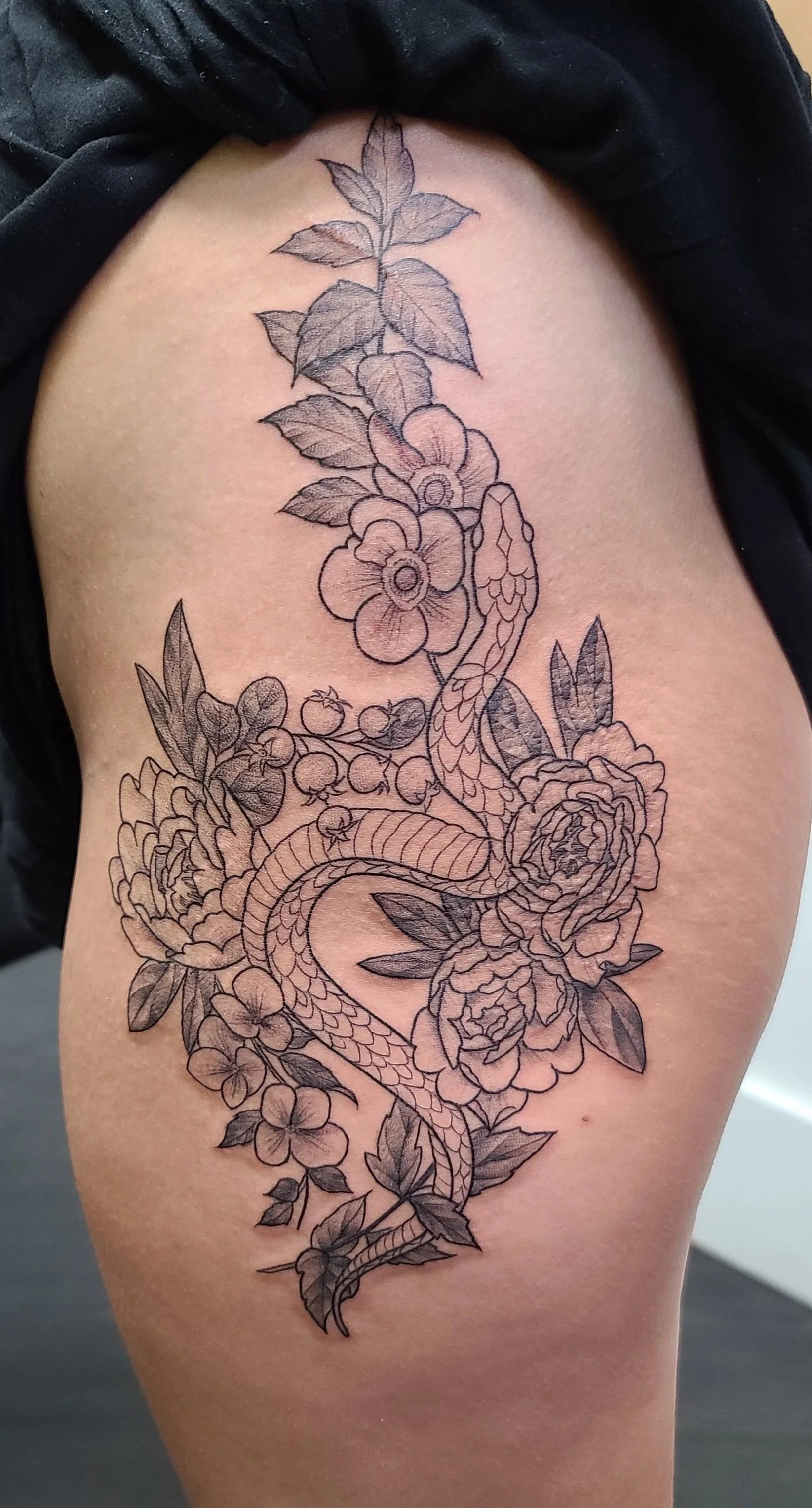 Line art tattoo of flowers, leaves, a snake, and a hummingbird on person's upper thigh.