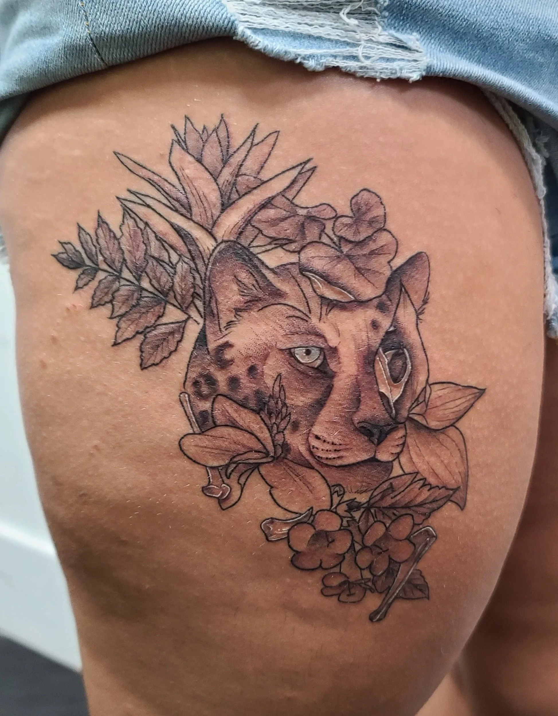 Tattoo of a cheetah's face with floral and plant designs on the thigh, in black and grey ink, with a mix of realistic and stylized elements.