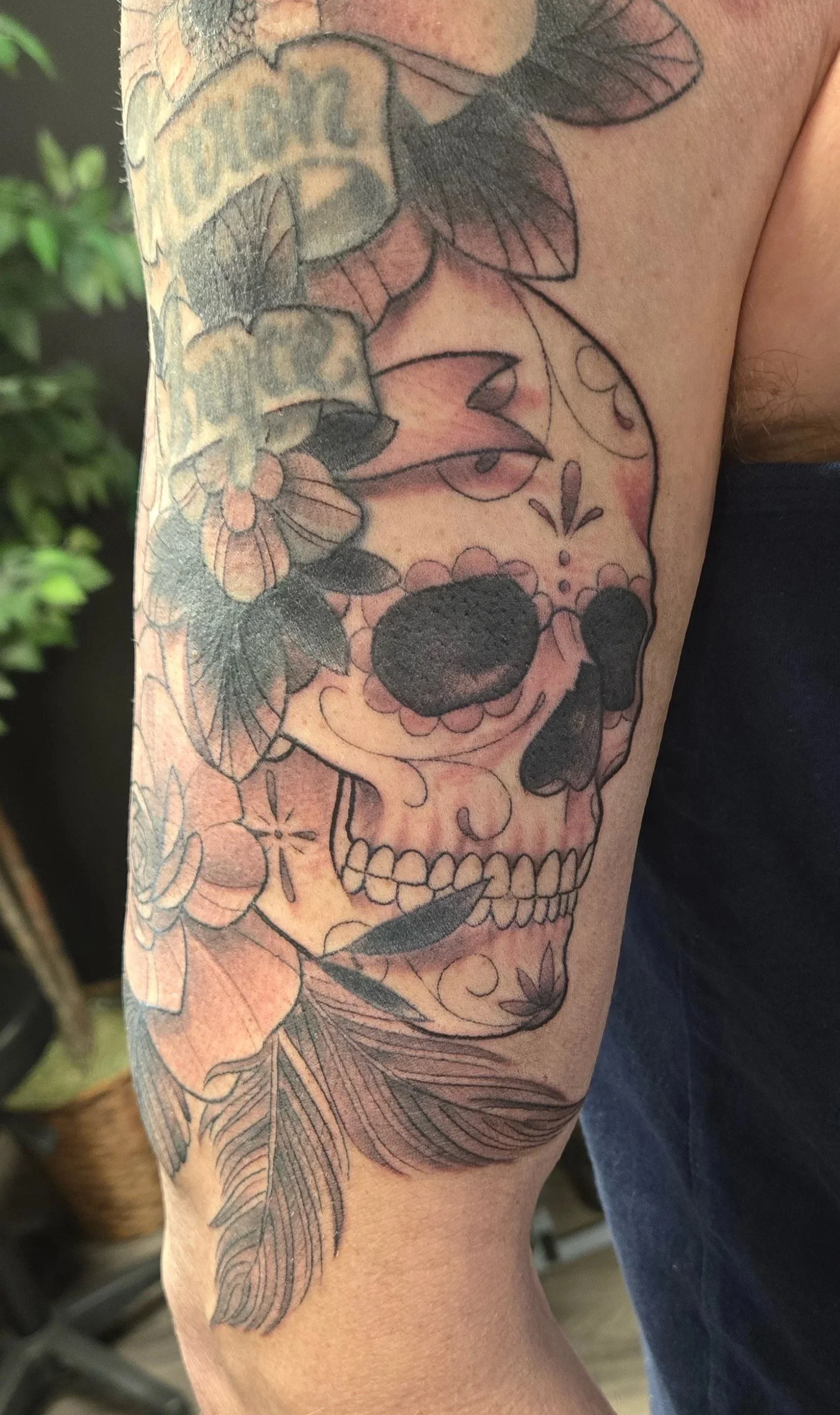 Tattoo of a sugar skull with floral and leaf accents on a person's upper arm.