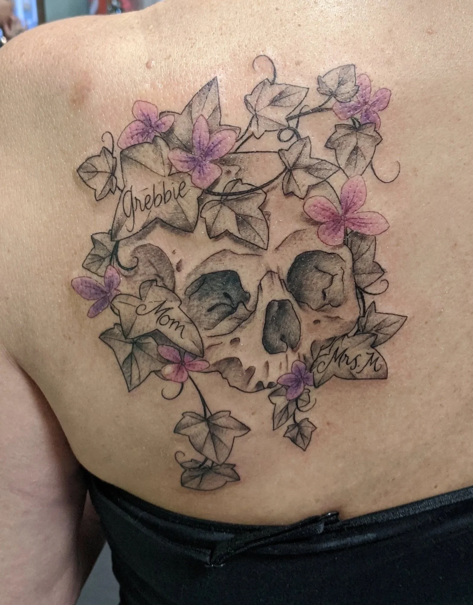 Tattoo of a skull surrounded by purple flowers and ivy on a person's shoulder blade, with the words 'Grebie', 'Mom', 'Mrs. K' written on the ivy.