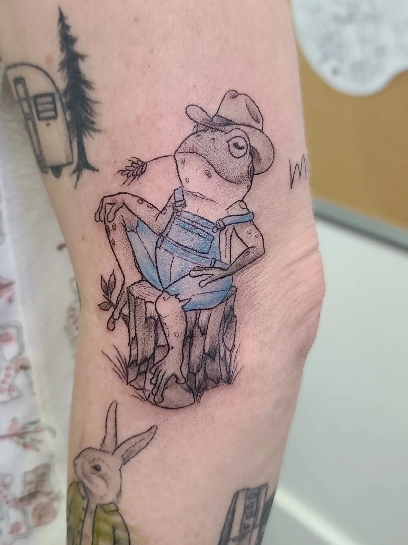 Tattoo of an anthropomorphic frog wearing overalls and a hat, sitting on a tree stump, with a rabbit standing nearby and a camper trailer in the background.