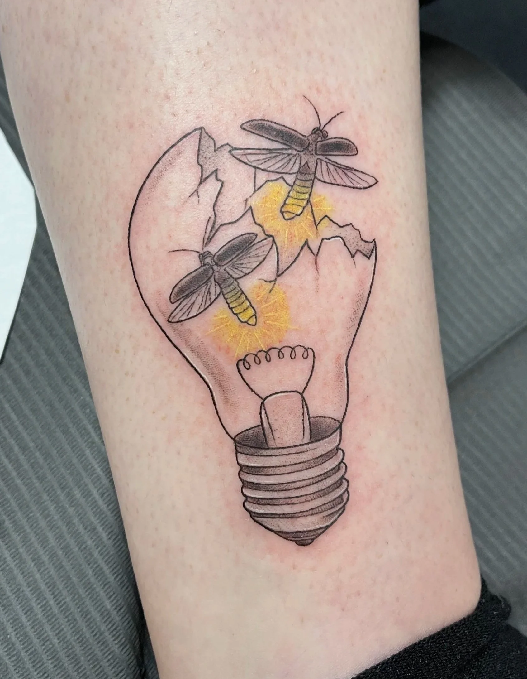 Tattoo of a broken light bulb with two bees flying around it on a person's arm.