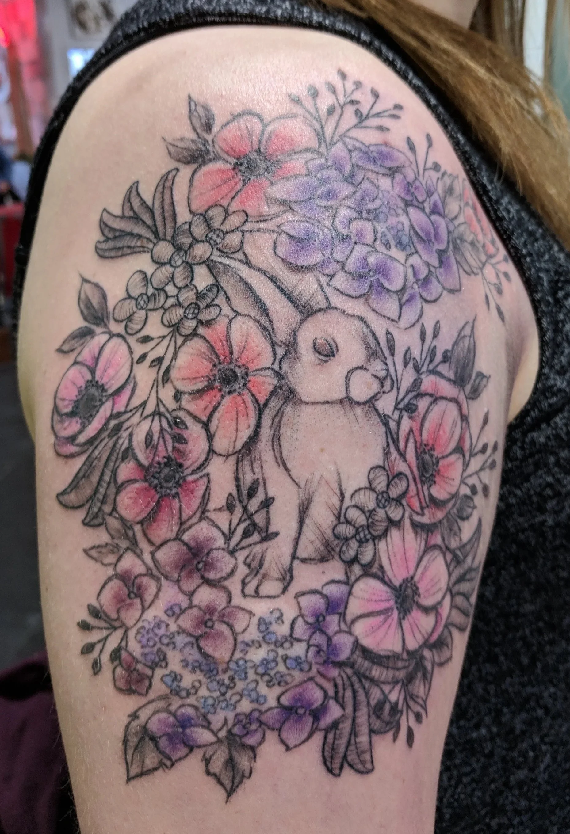 Tattoo of a bunny surrounded by pink and purple flowers on upper arm.