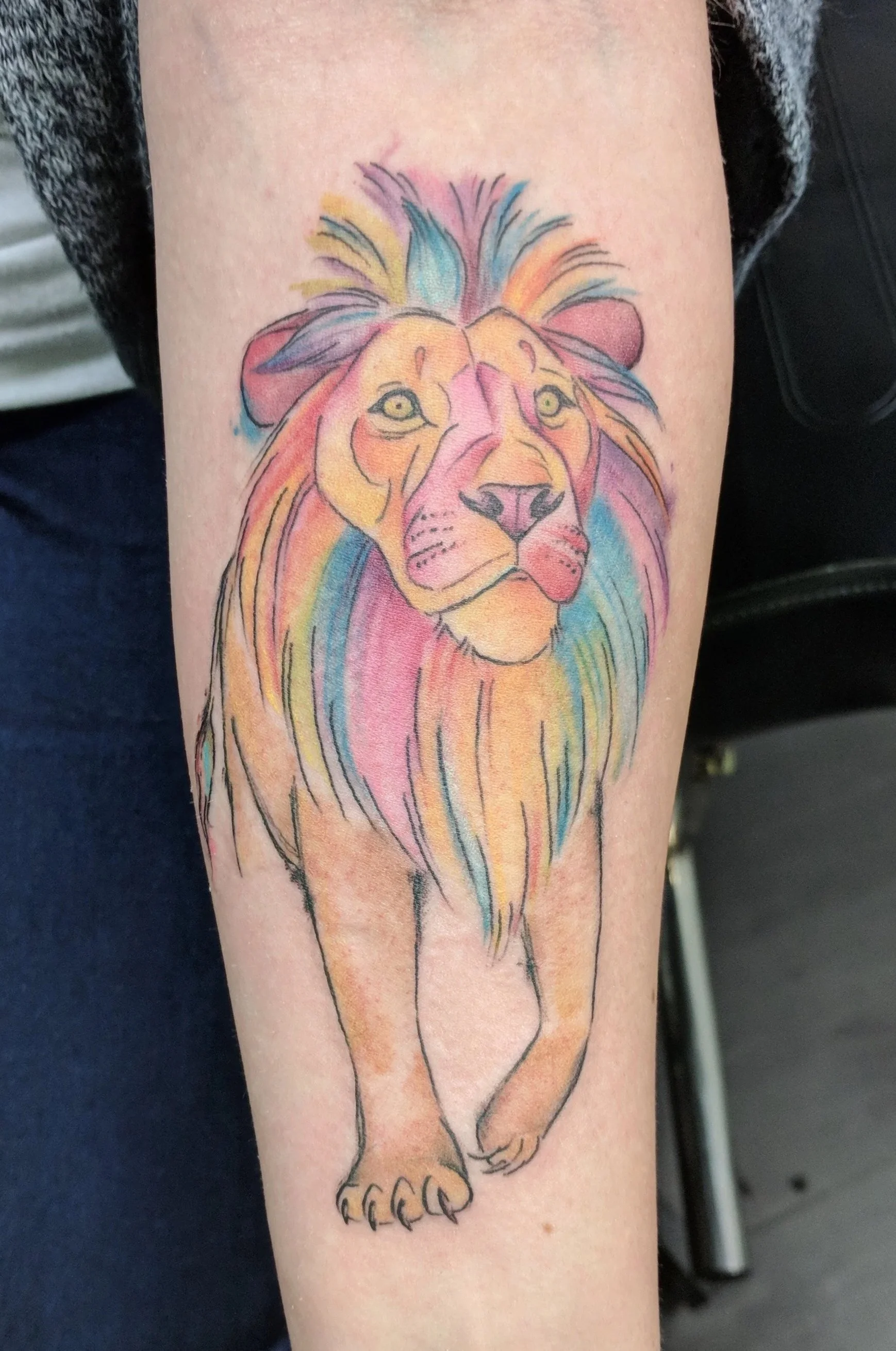 Watercolor-style tattoo of a lion on a person's forearm, featuring a colorful mane and face with shades of pink, purple, blue, yellow, and orange.