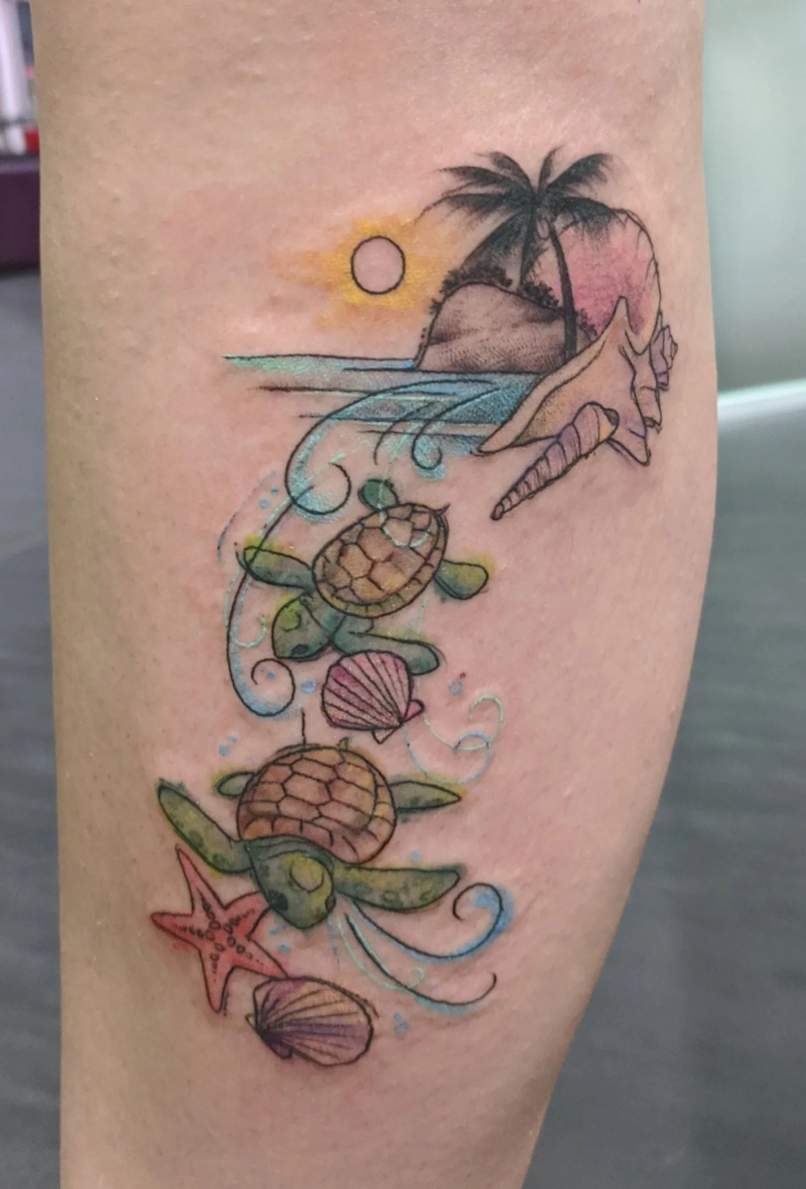 Colorful tattoo of a beach scene with palm trees, a sunset, a unicorn, turtles, shells, a starfish, and waves.