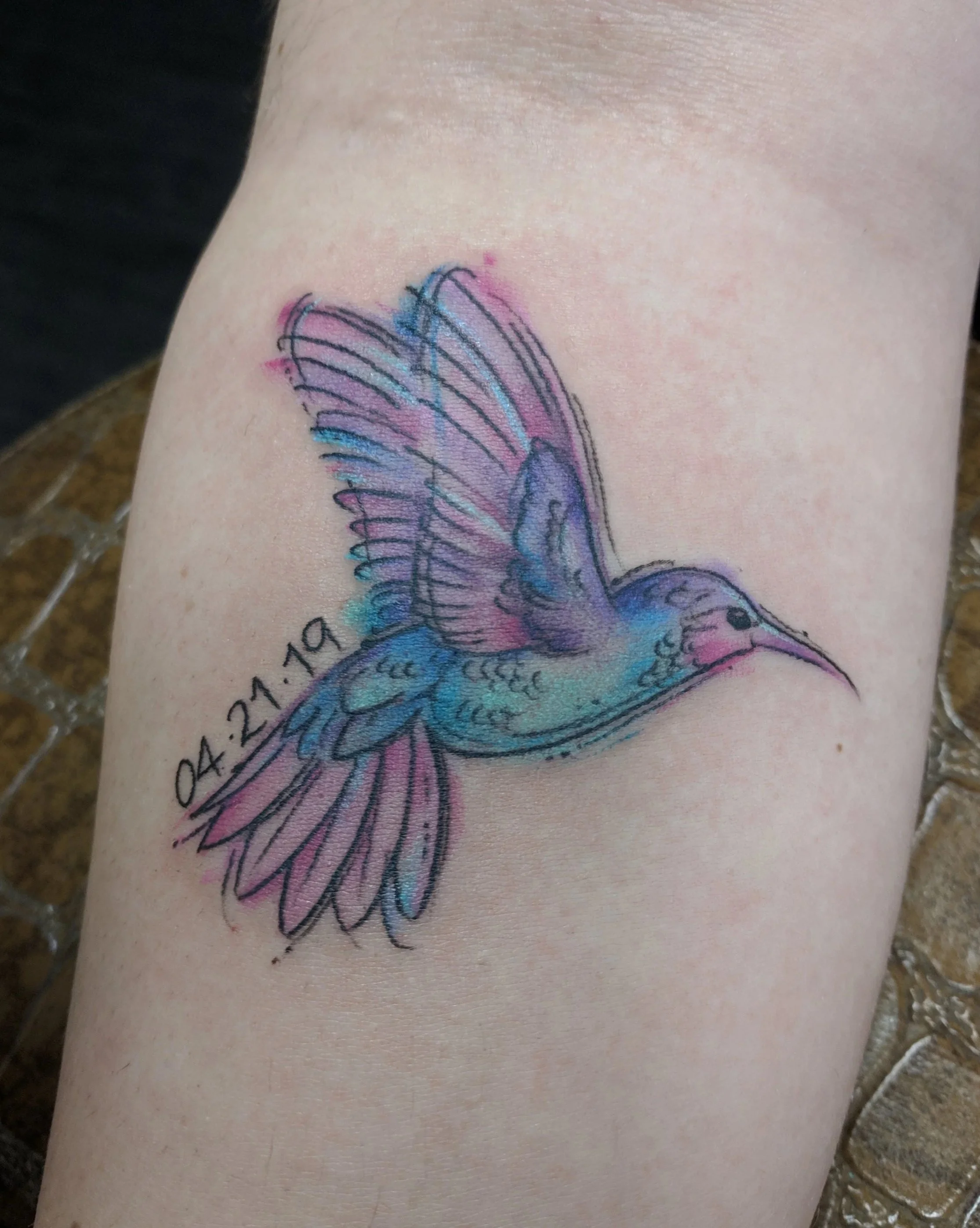 Colorful tattoo of a hummingbird with outstretched wings and the date '04.21.19' on the skin.