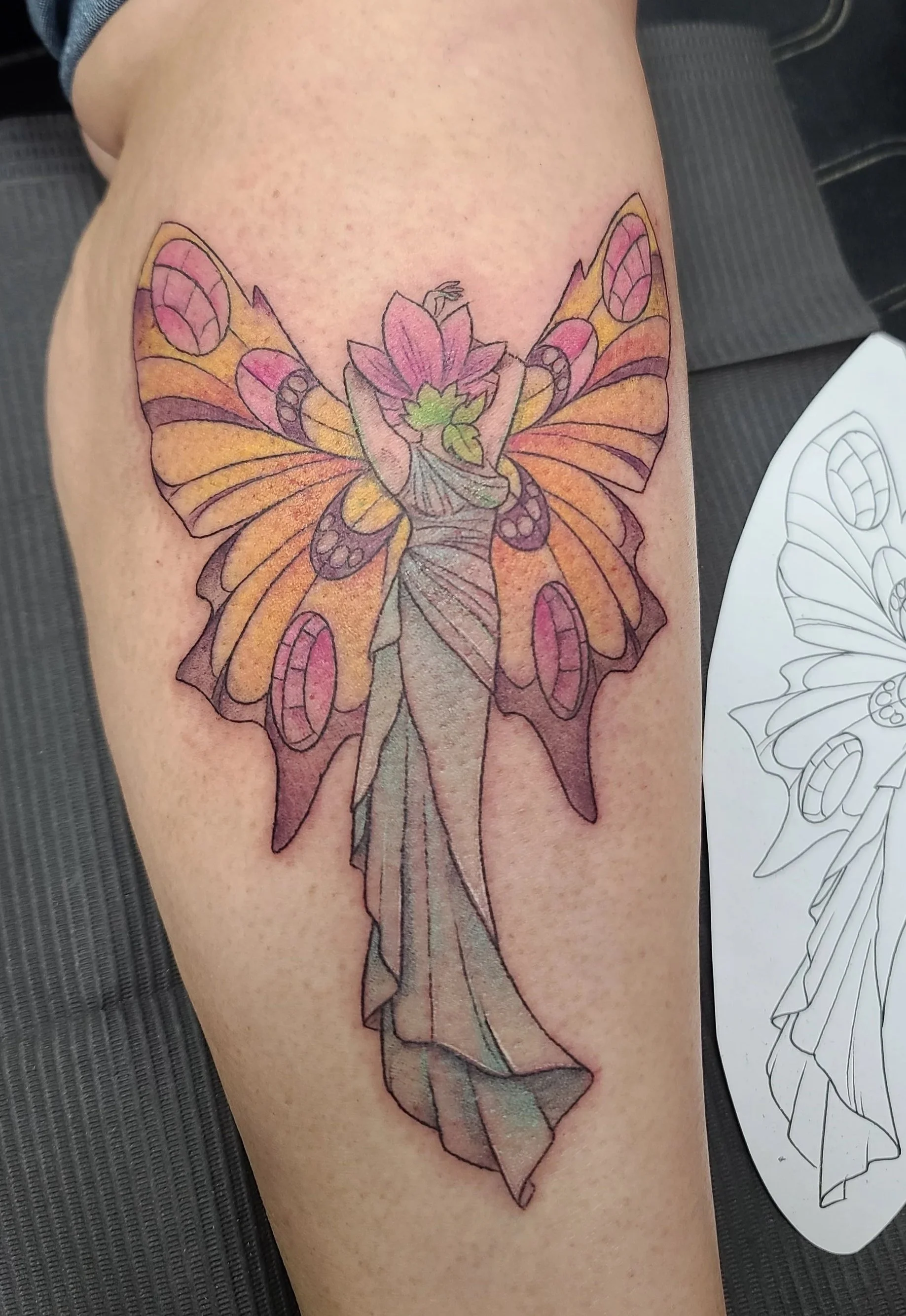Colorful tattoo of a fairy with butterfly wings and a flower in her hair on a person's thigh, with a black and white sketch of the design next to it.