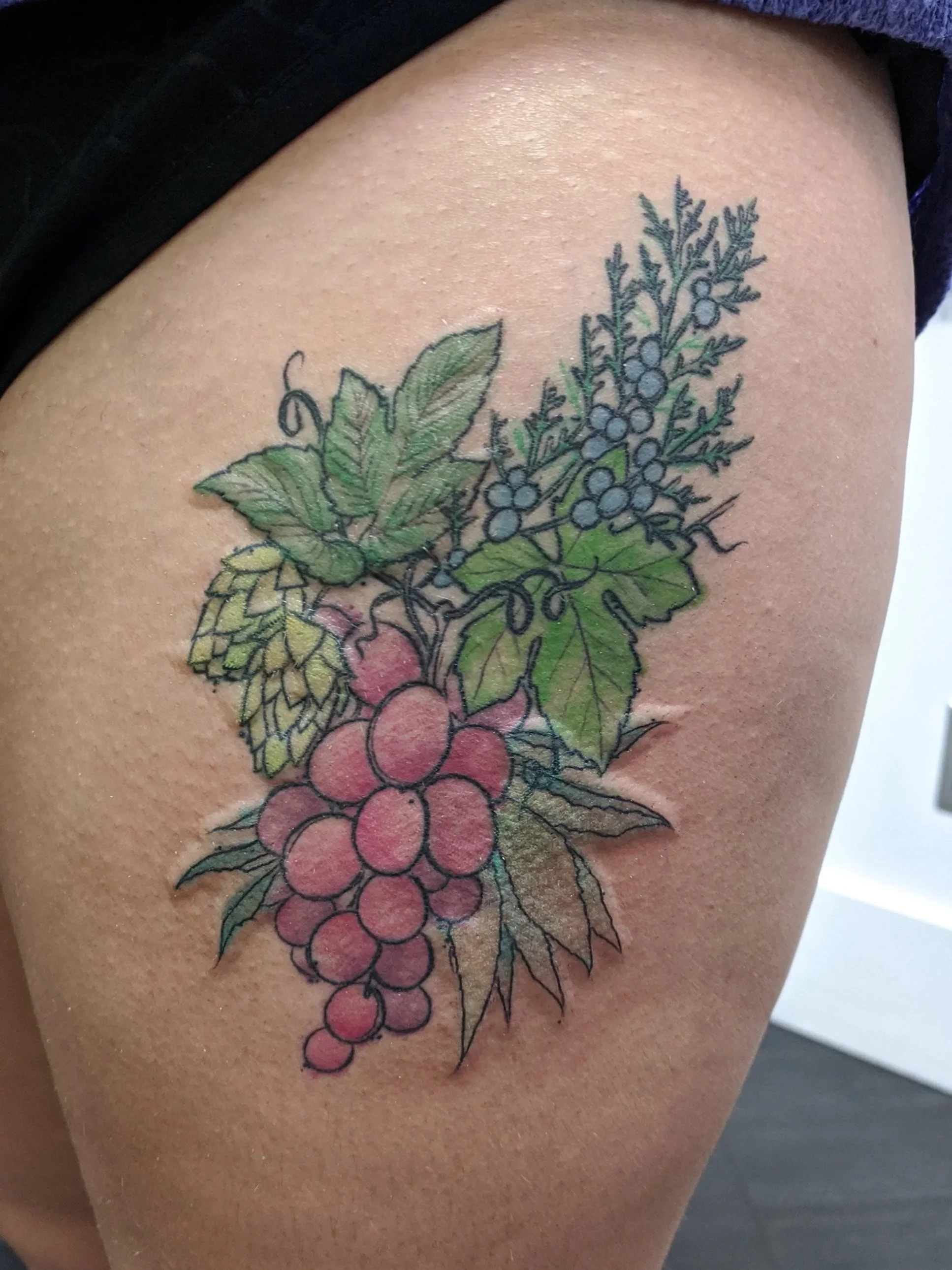 Colorful tattoo of a grapevine with green leaves, purple grapes, and pinkish-red grapes on a person's thigh.