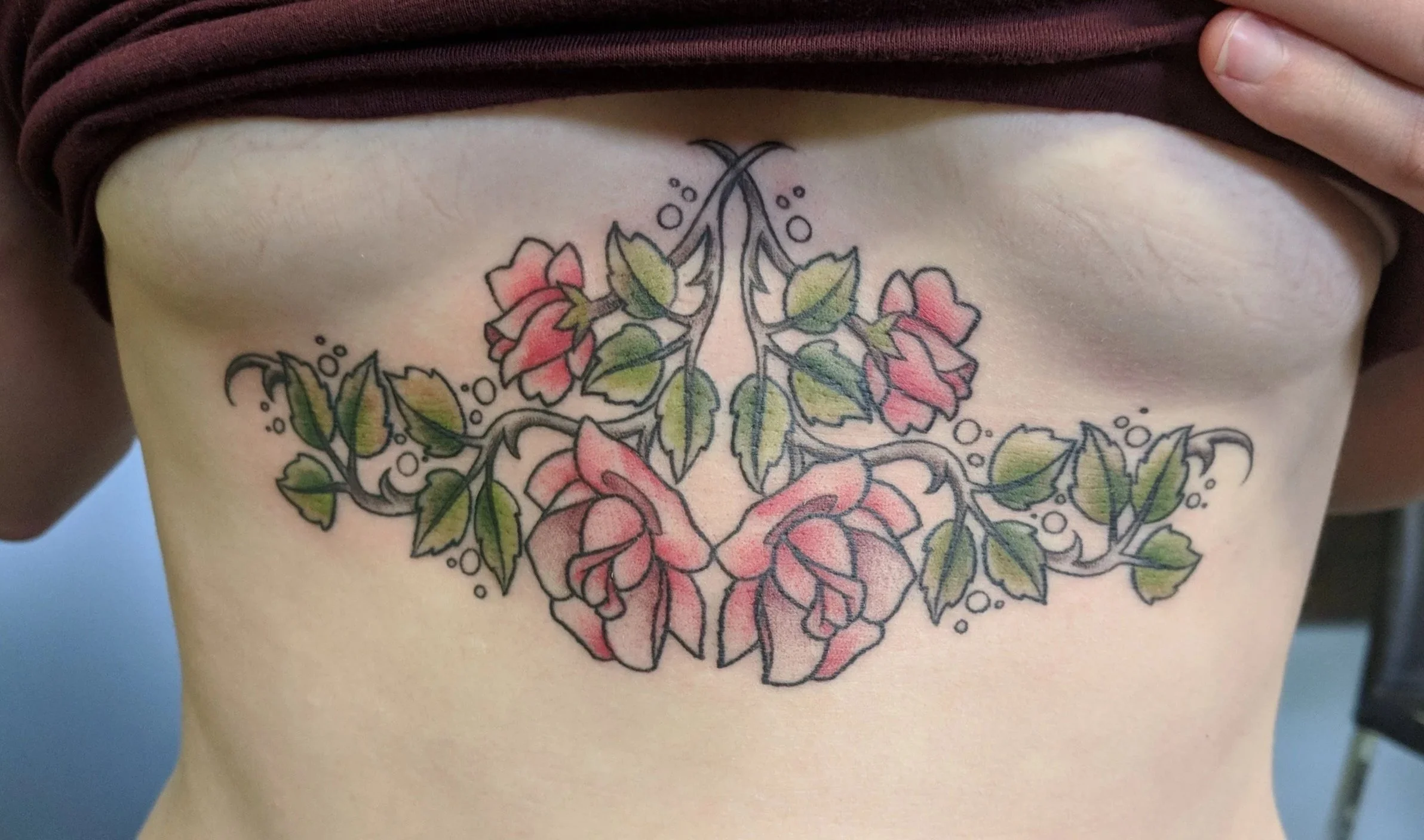A tattoo of pink roses with green leaves and black outlines on the person's lower ribcage.