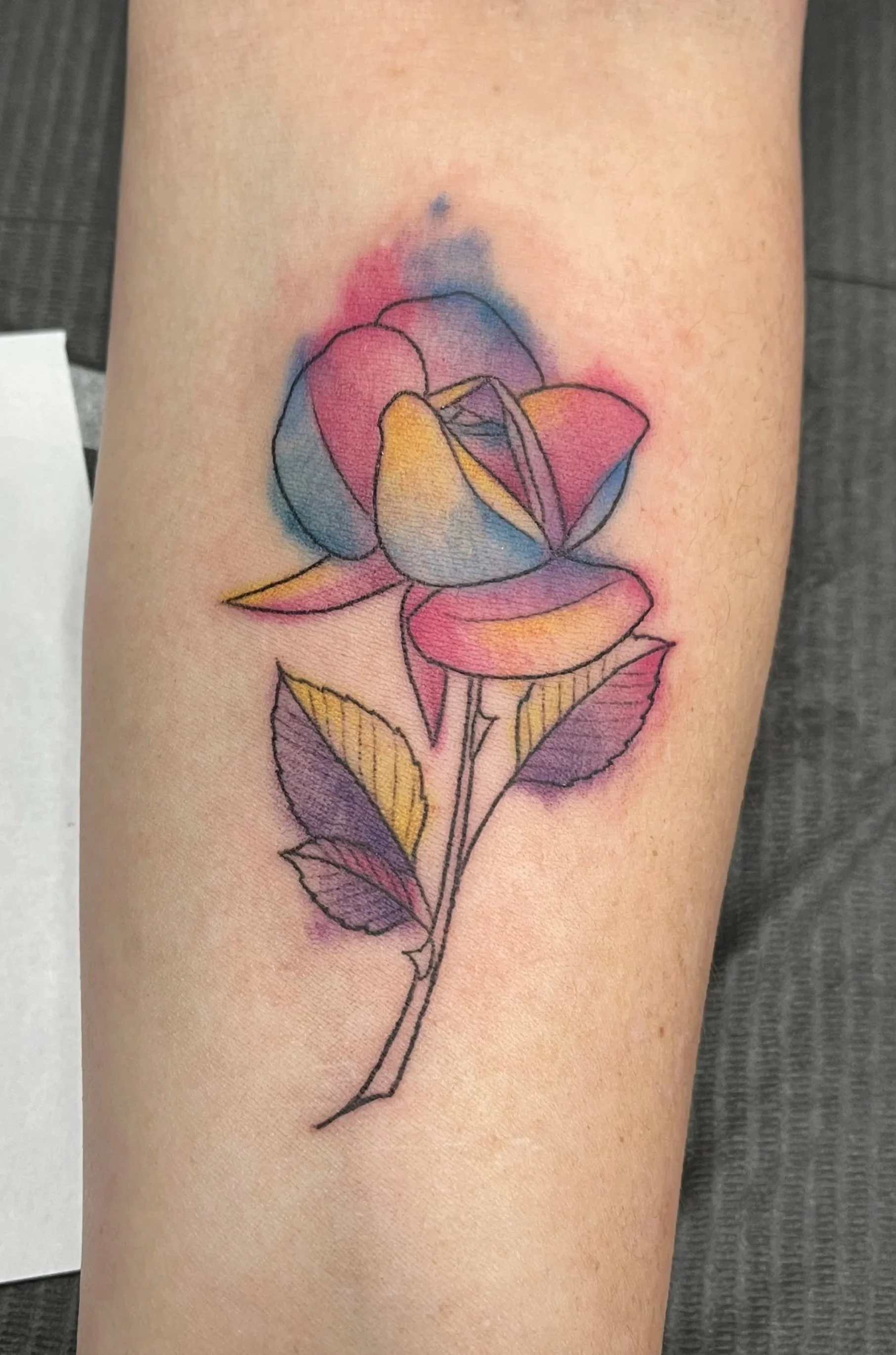 Watercolor-style tattoo of a rose with pink, yellow, and purple petals, green and purple leaves, on a person's forearm.