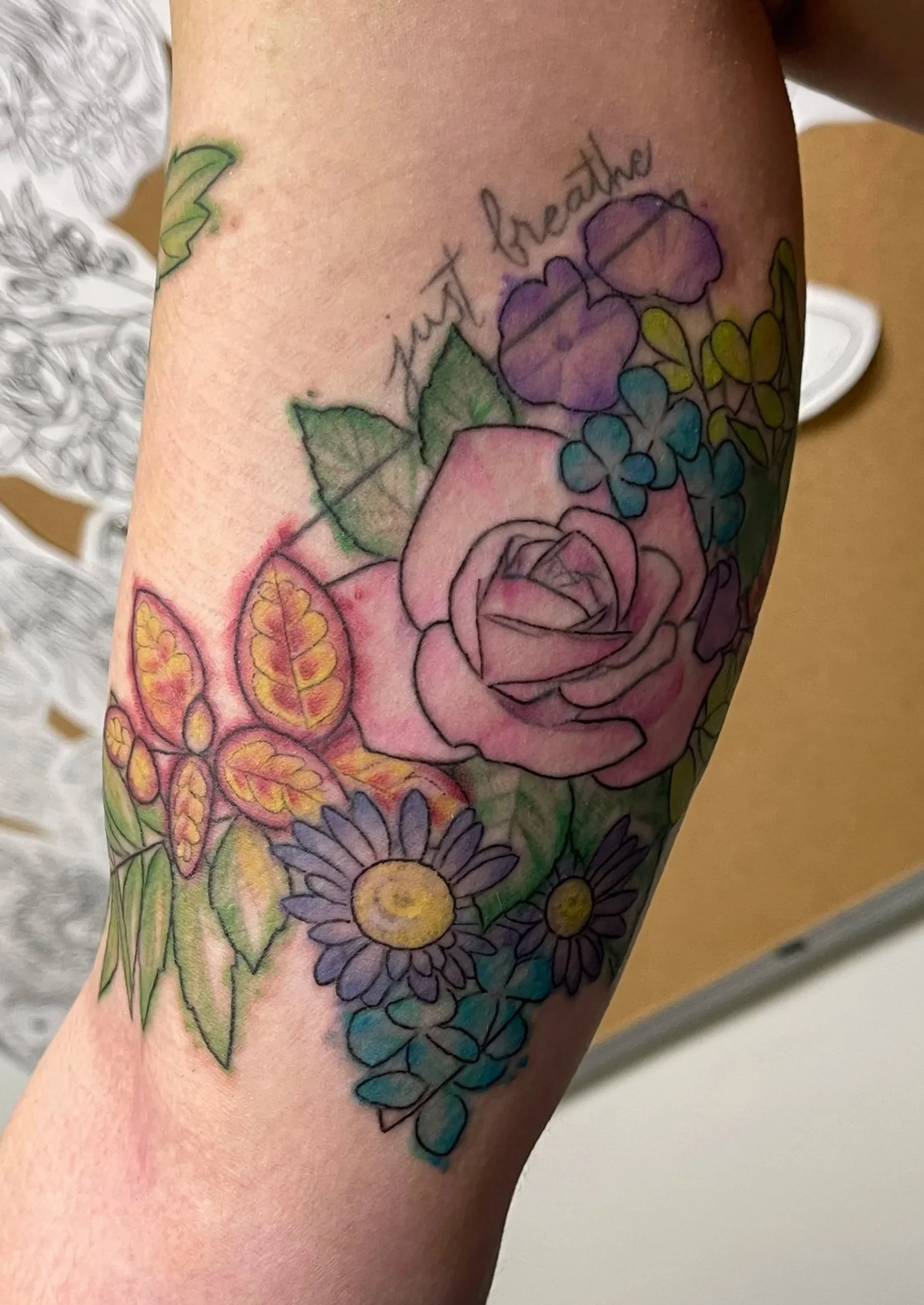 Colorful floral tattoo on a person's arm featuring a pink rose, purple and blue flowers, yellow and purple leaves, and the phrase 'just breathe' written above the flowers.
