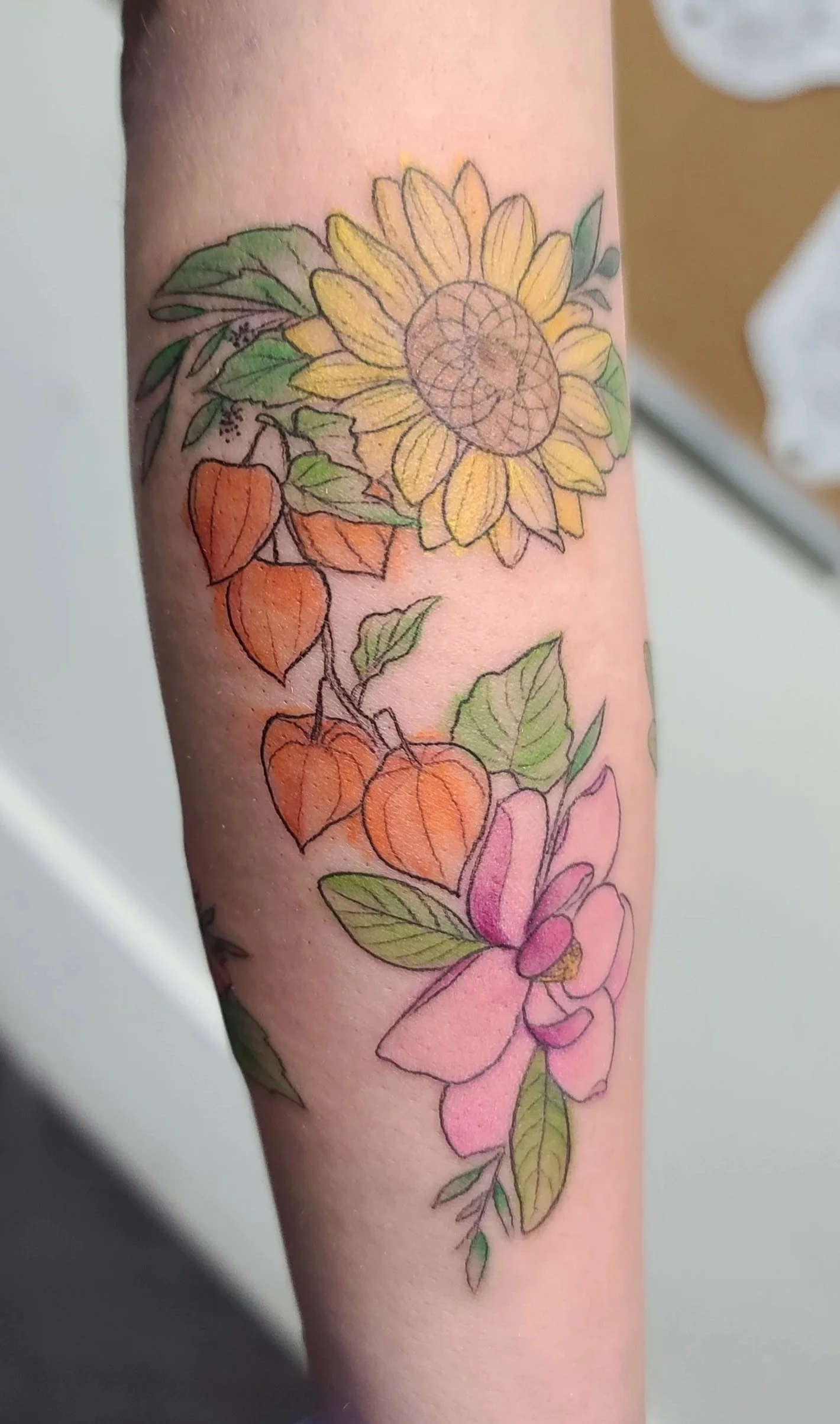 Colorful tattoo of a sunflower, orange lantern plants, and pink flowers on person's forearm.