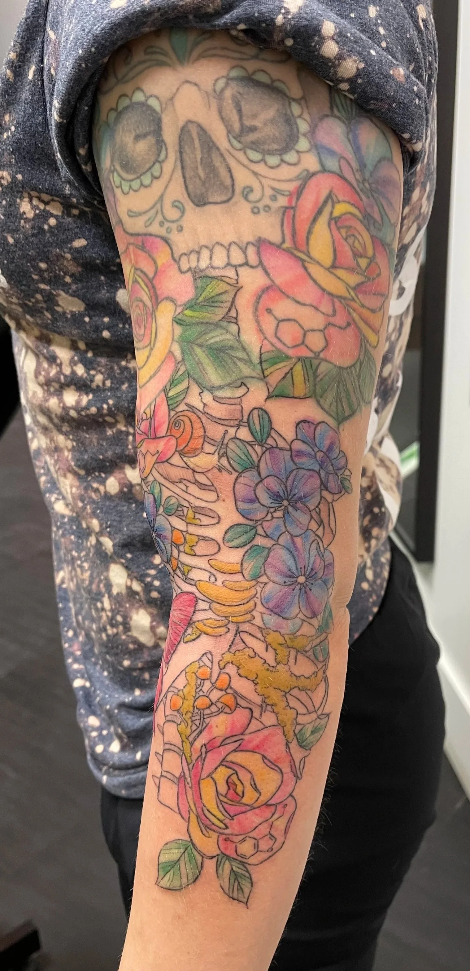 Colorful tattoo of a skull, roses, and flowers on a person's upper arm.