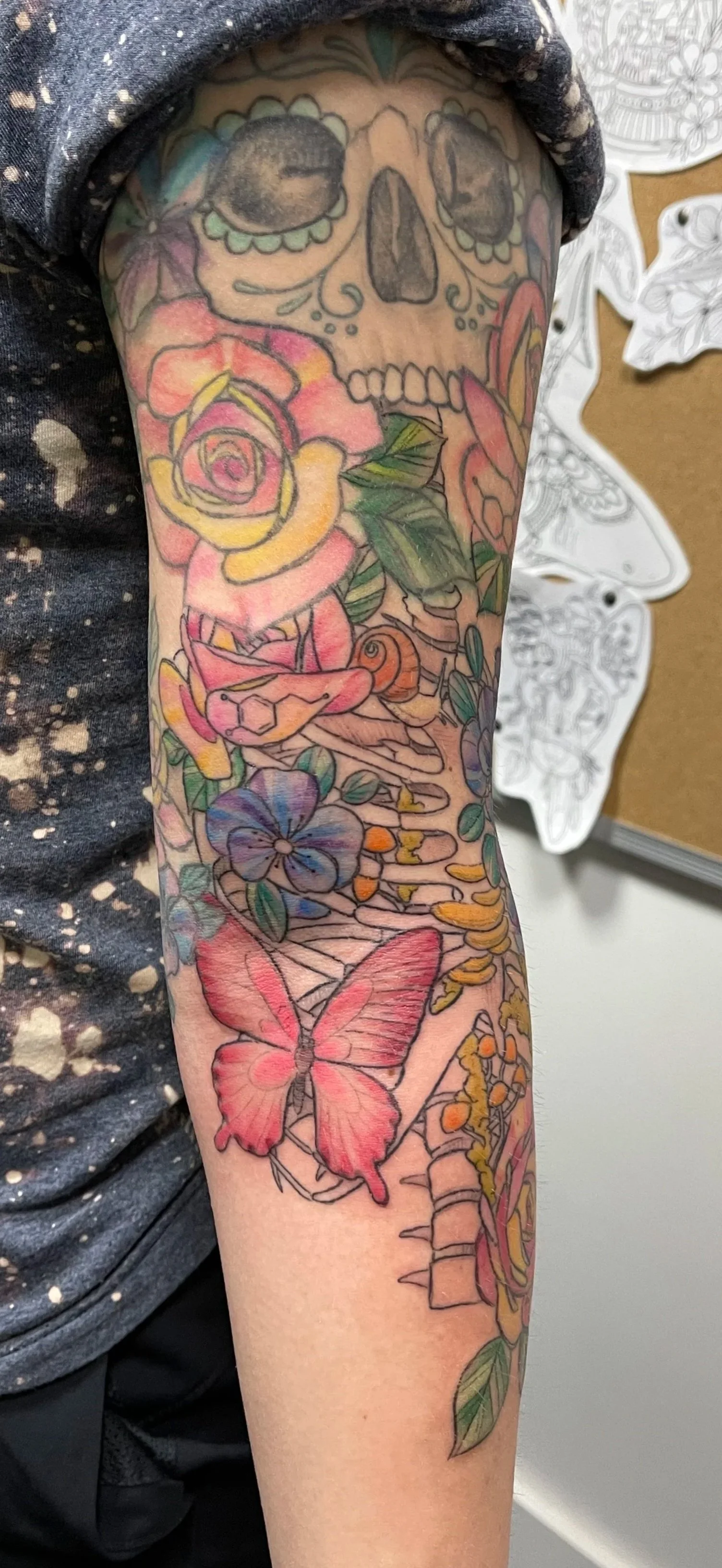 Colorful tattoo on person's arm featuring a skull, roses, a butterfly, flowers, and other decorative elements.