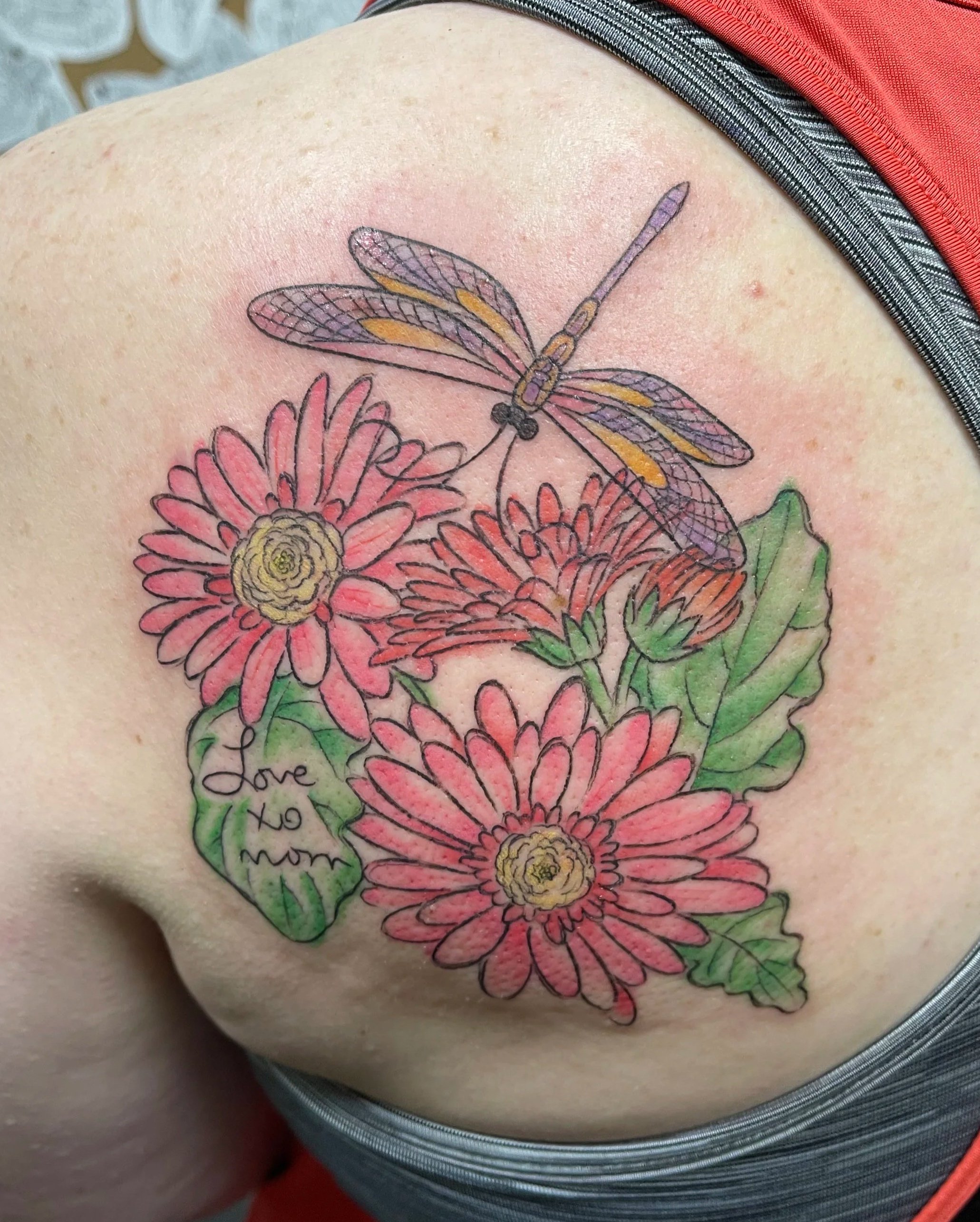 Colorful tattoo of pink and yellow flowers with green leaves and a purple and yellow dragonfly, with the words "love to mom" written on one leaf, on a person's shoulder.