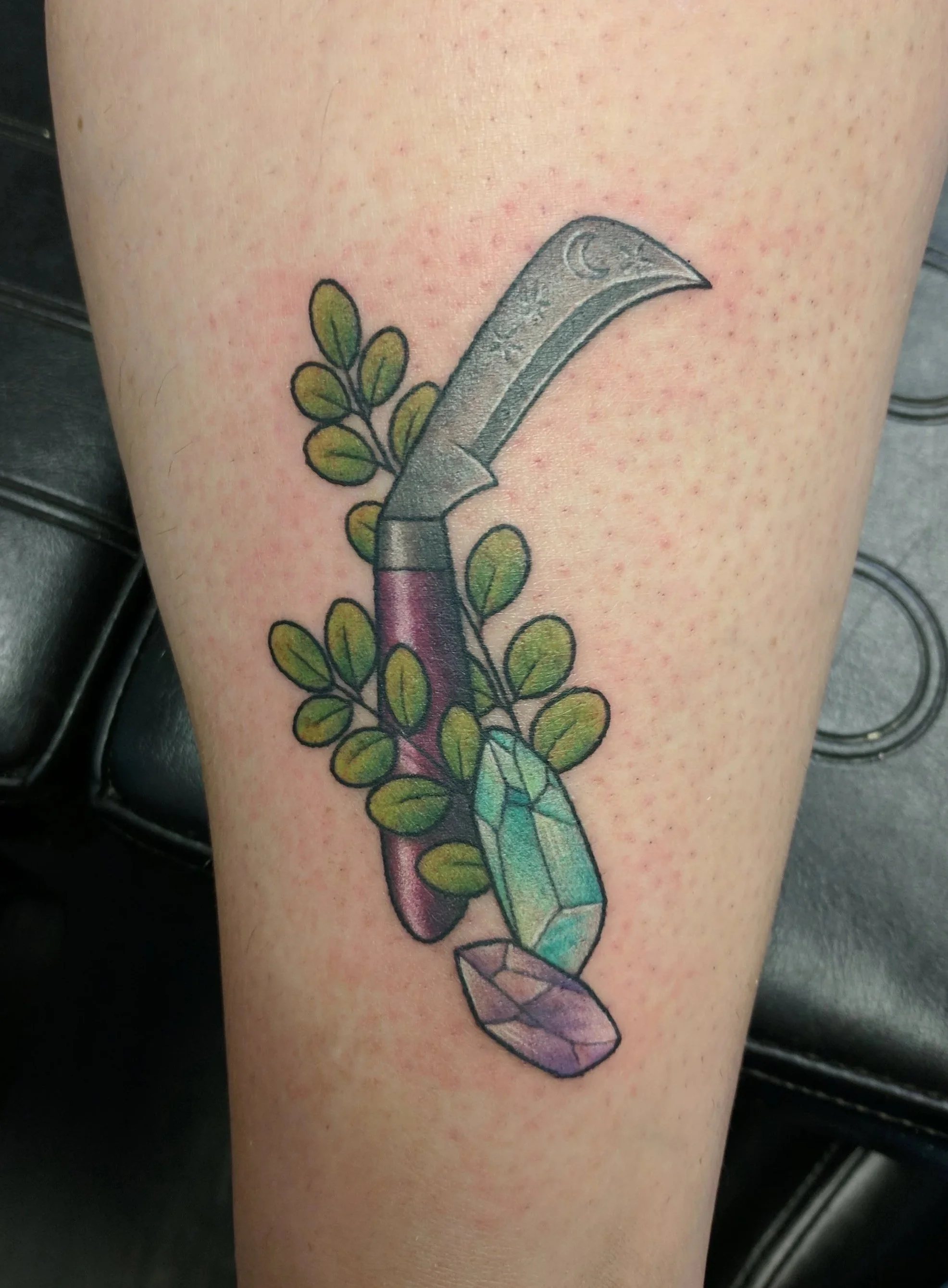 A tattoo of a gray knife with green leaves around it, and two crystals at the bottom, one purple and one blue-green.