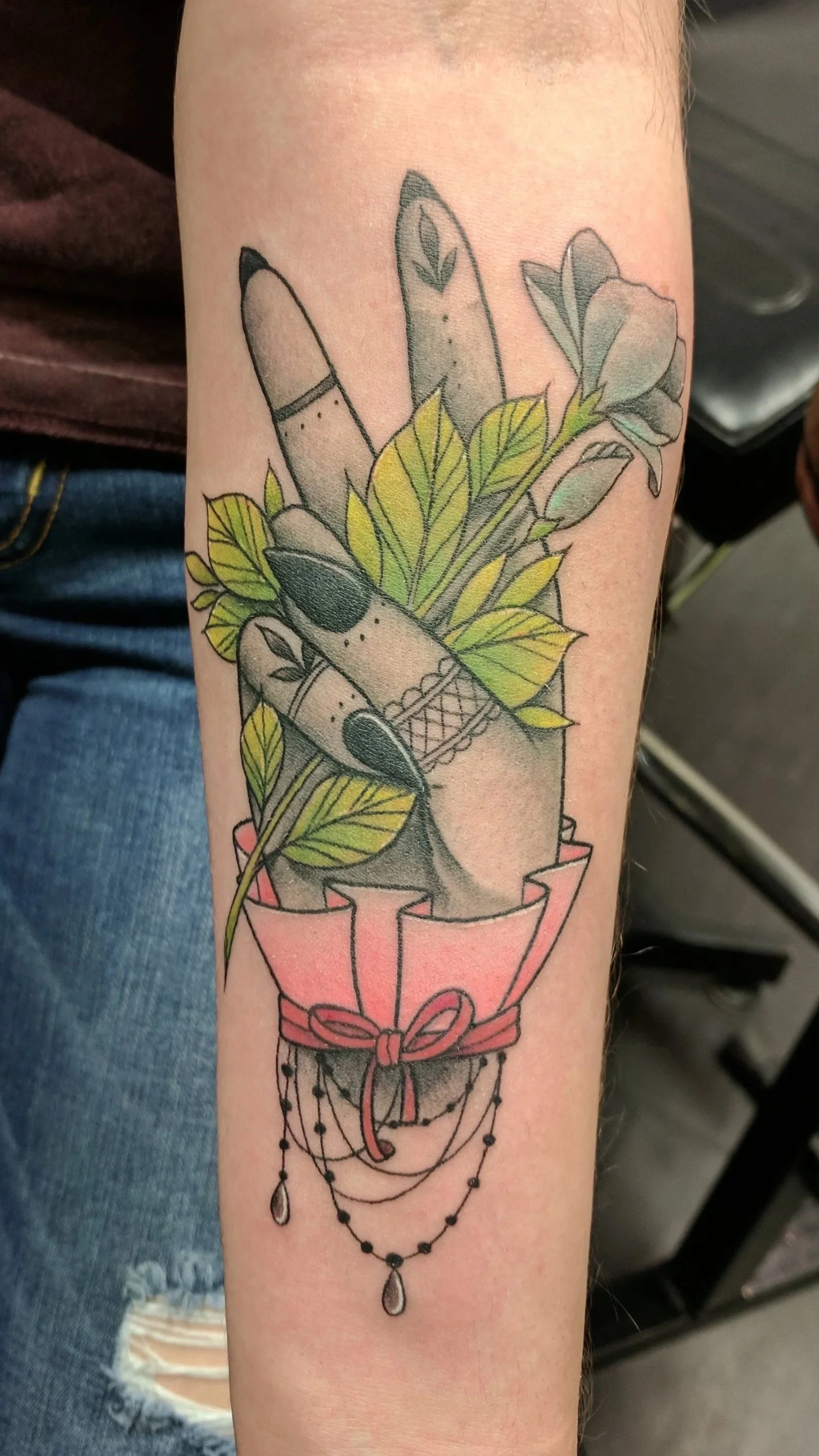 Tattoo of a hand with decorated fingers, holding a bouquet of green leaves, inside a pink wrapper with hanging beads.