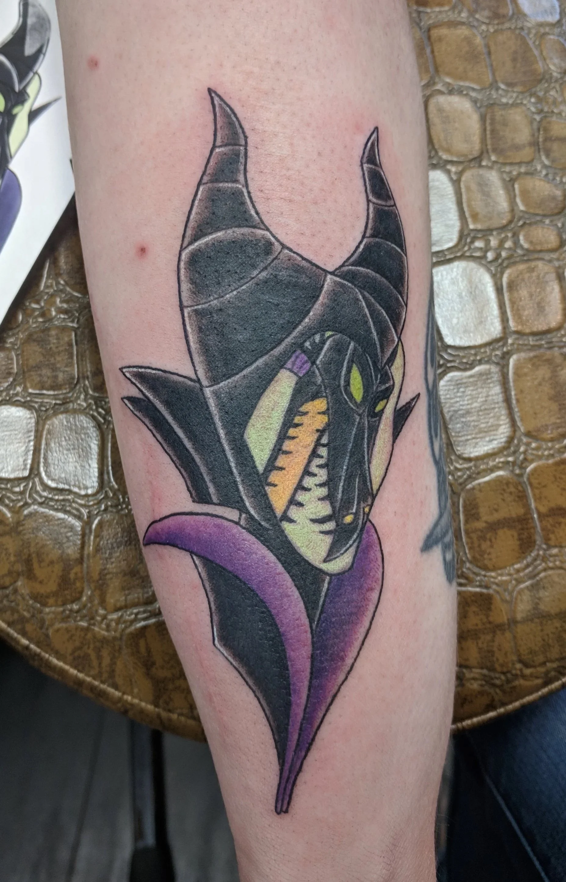 A colorful tattoo of a black cat with yellow eyes, purple tongue, and green highlights on the cheek, styled in a traditional tattoo design on a person's forearm.