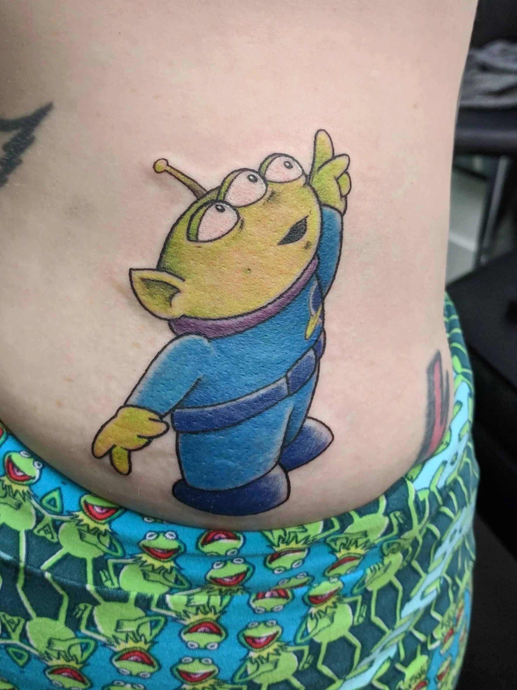 Tattoo of a green alien with three eyes, wearing a blue jumpsuit, and pointing upward, on a person's side.
