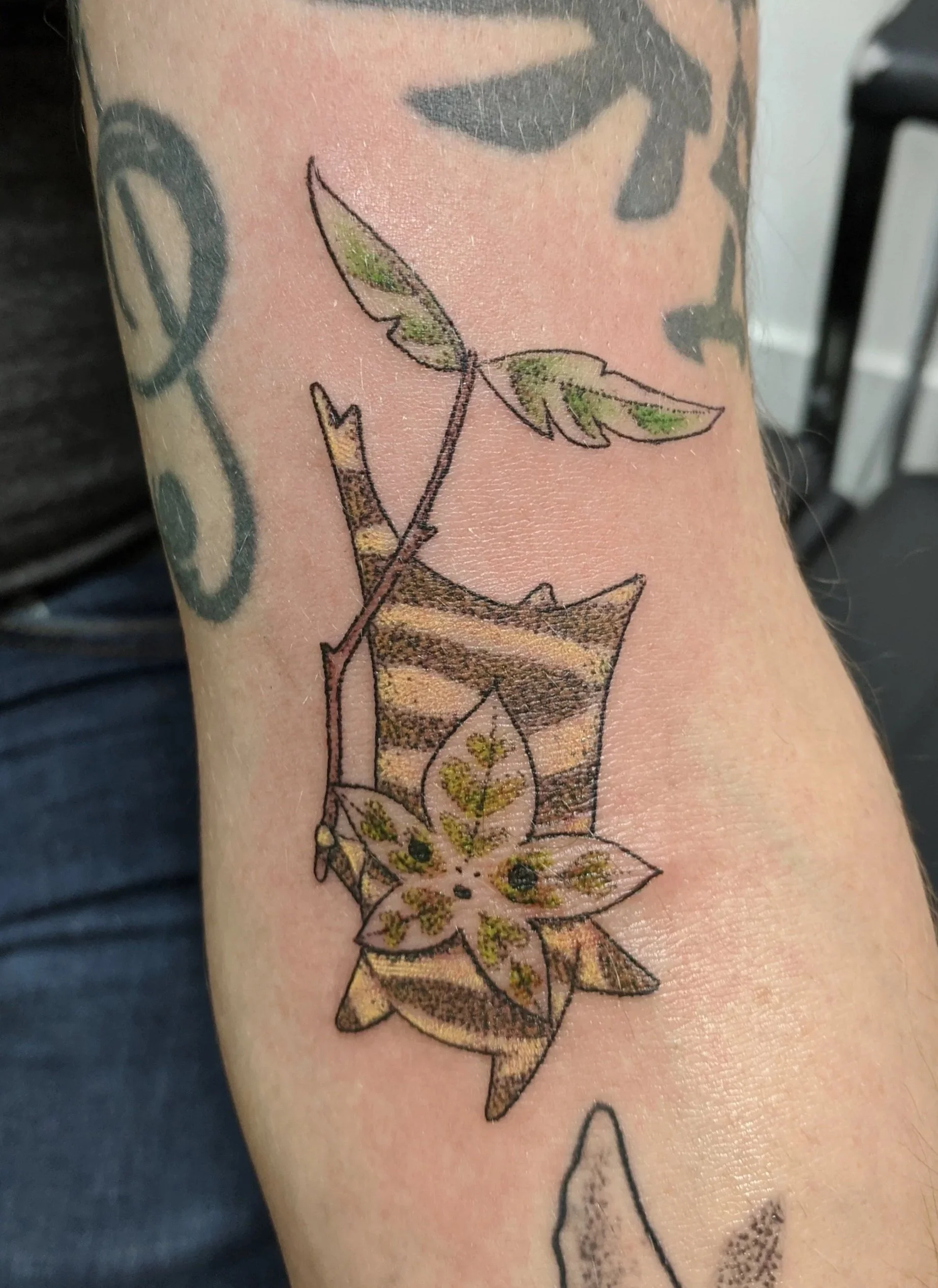 Tattoo of a branch with green leaves and a flower, with a background pattern of brown and black stripes.