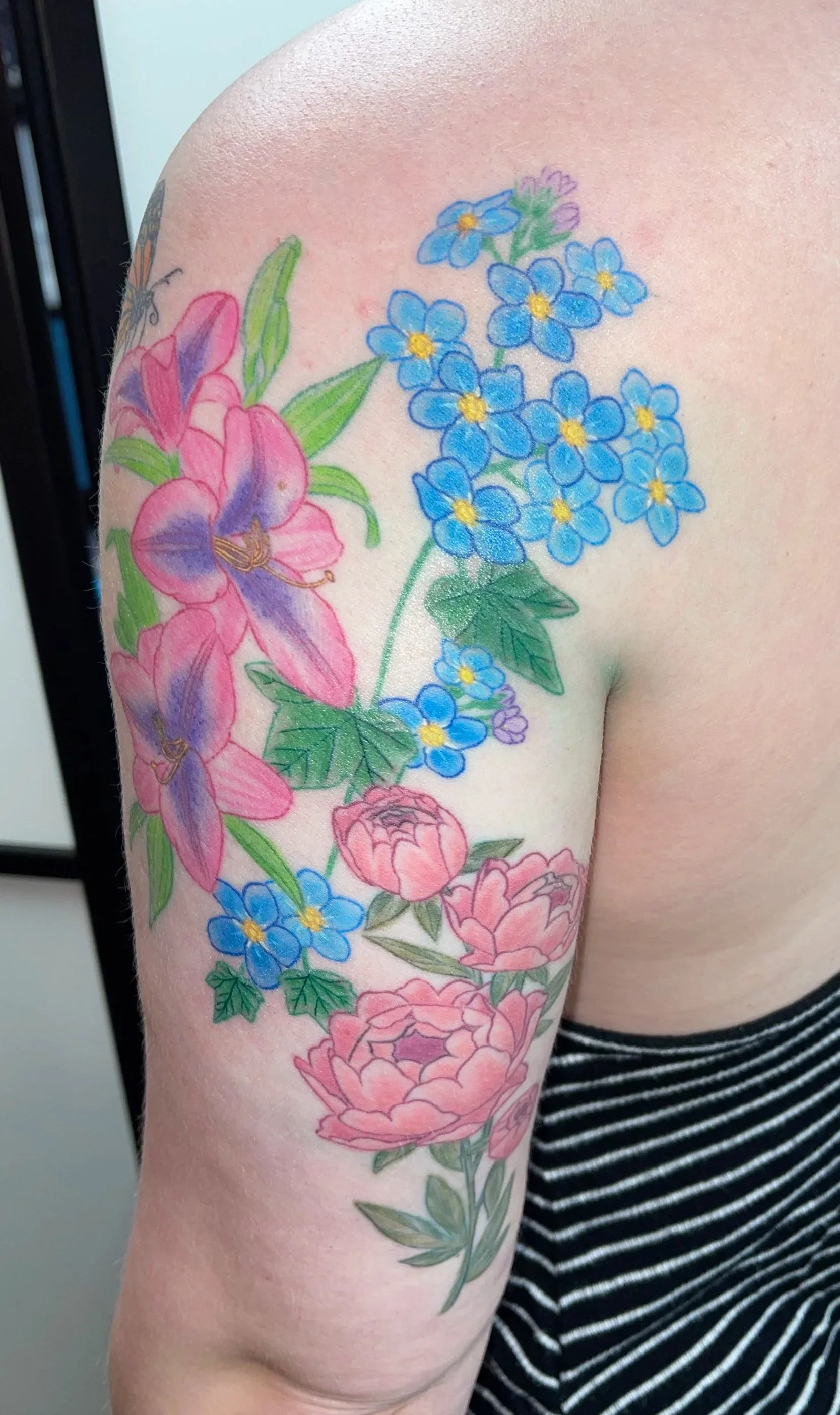 A colorful floral tattoo on a person's shoulder and upper arm, featuring pink and purple flowers, blue flowers, and green leaves.