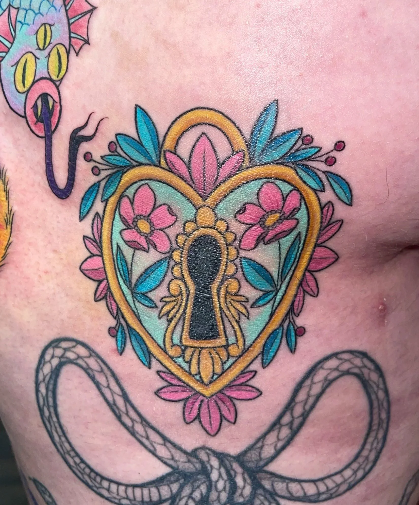 Colorful tattoo of a heart-shaped lock surrounded by pink and blue flowers on a person's thigh, with tattoos of a snake and other designs visible.