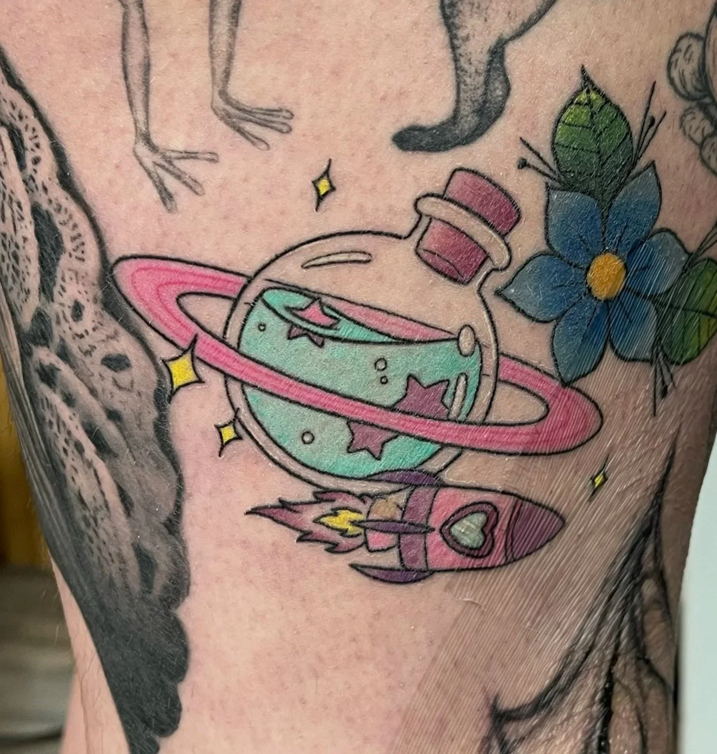 Tattoo on person's leg featuring a frog, a fox wearing clothes, a flower, a rocket, and a spaceship with a planet.