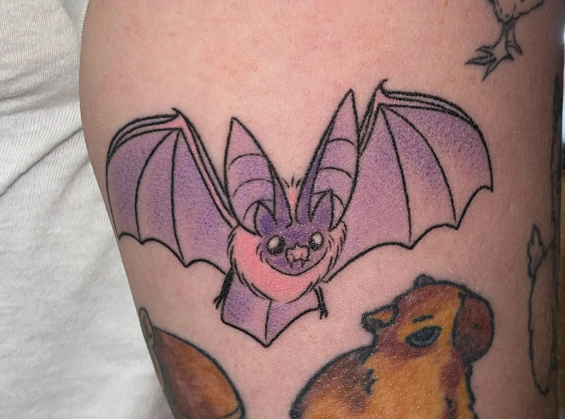 Tattoo of a purple bat with large wings and small body on person's upper arm, surrounded by tattoos of a chicken, a dog, and a butterfly.