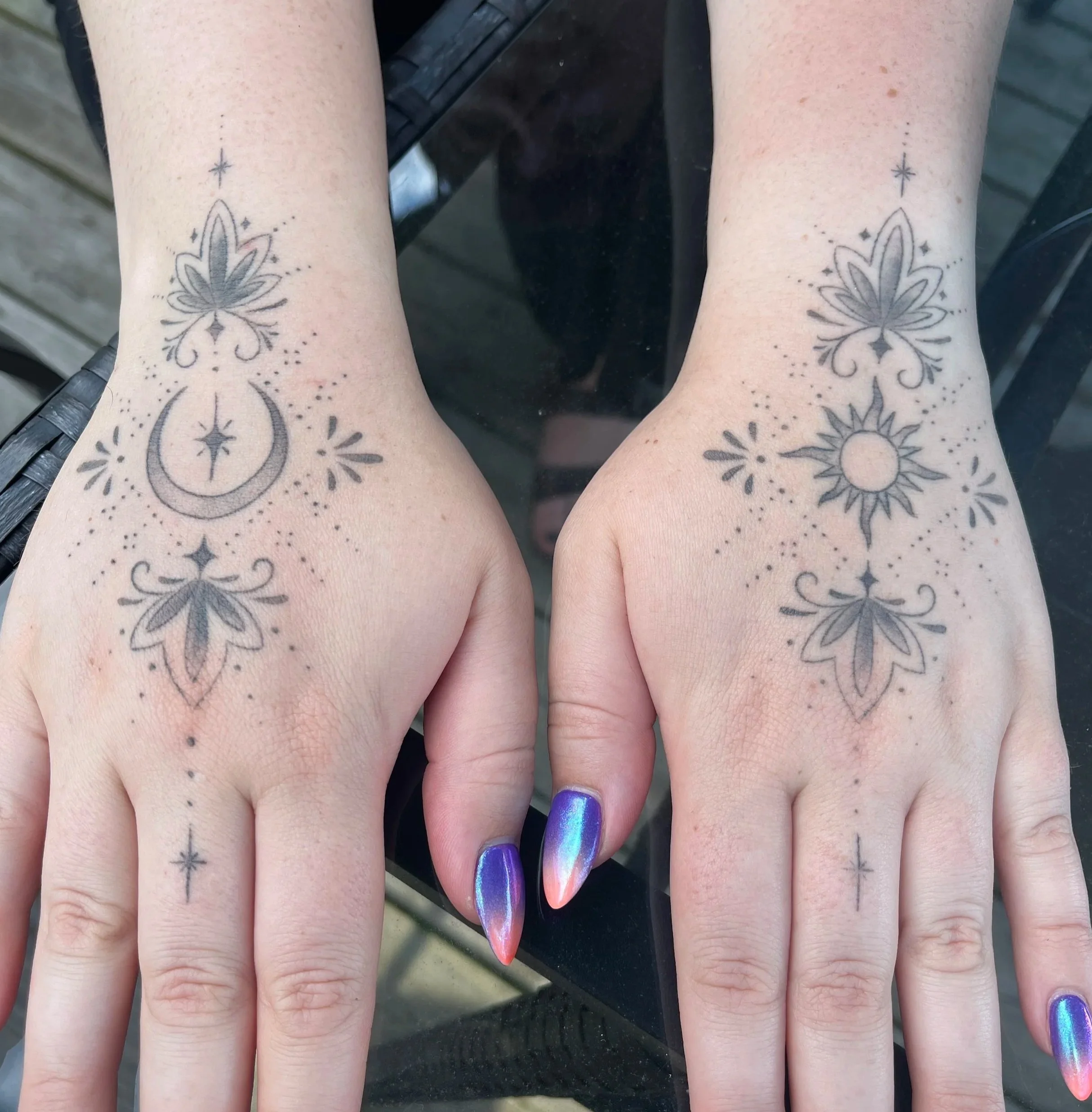 Matching tattoos on both hands featuring celestial and floral designs, including a moon with a star, a sun, and decorative elements, with metallic gradient nails in purple and pink.
