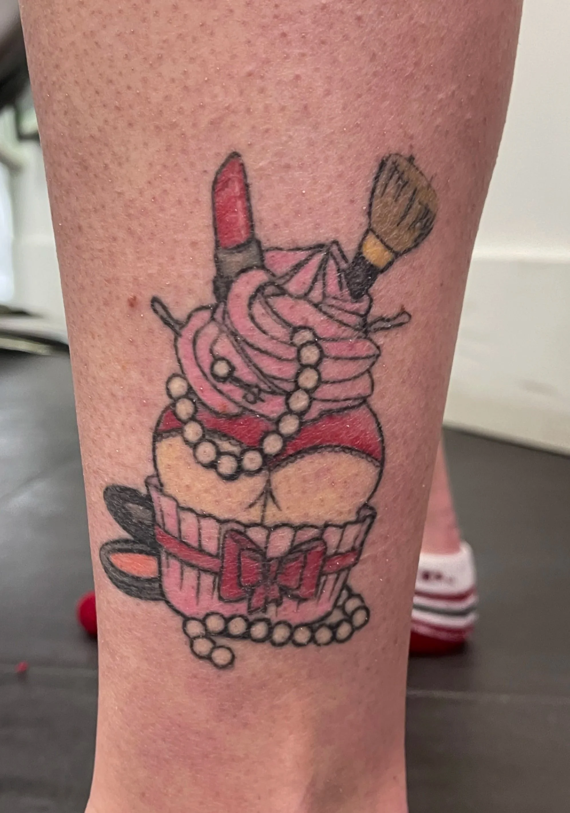Tattoo of makeup and jewelry from a pink cupcake with a cherry on top, decorated with a pearl necklace, lipstick, makeup brush, and bow.