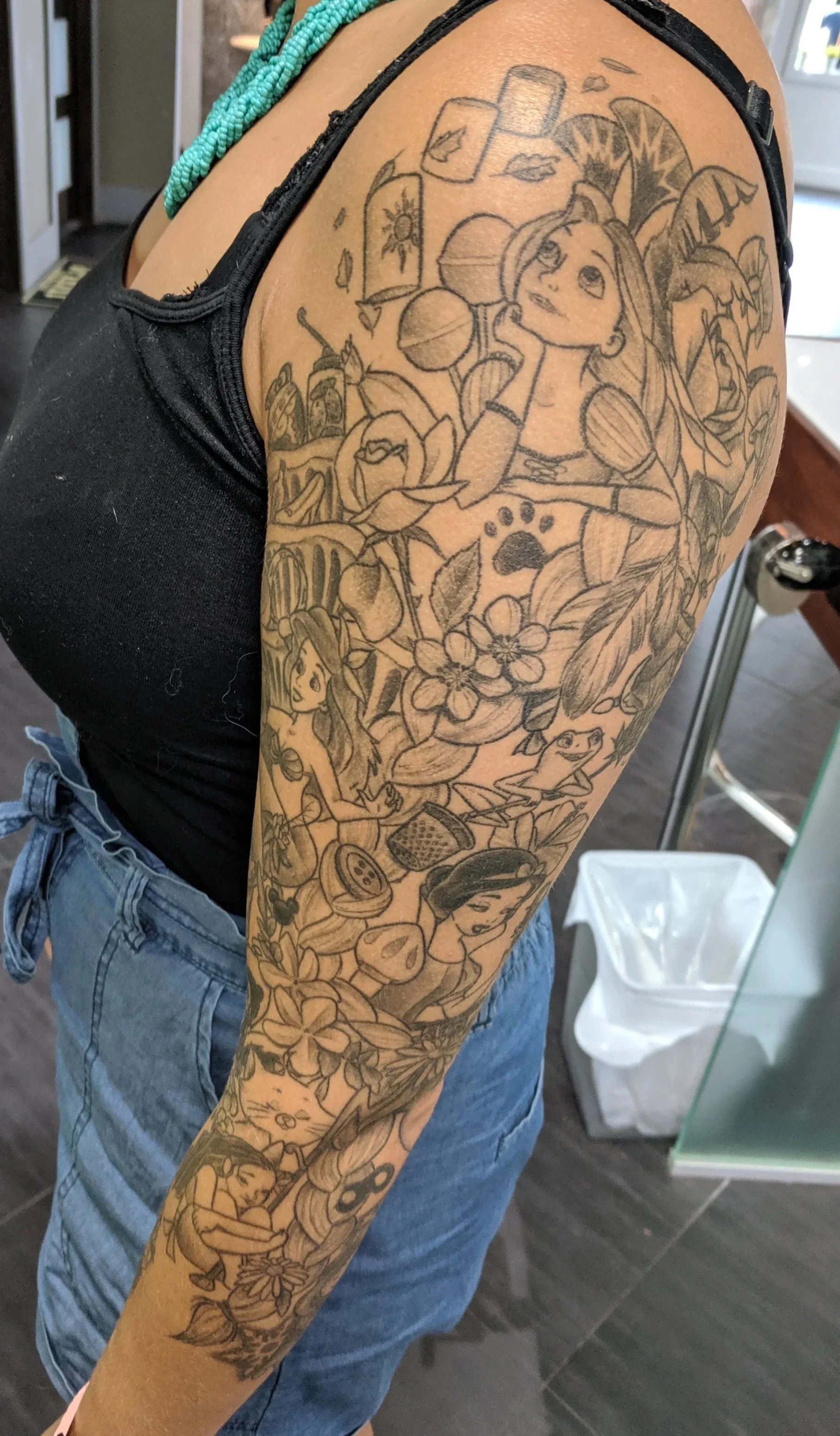 A detailed black and gray sleeve tattoo on a woman's arm, featuring a mermaid, flowers, a paw print, a girl, and various small objects.