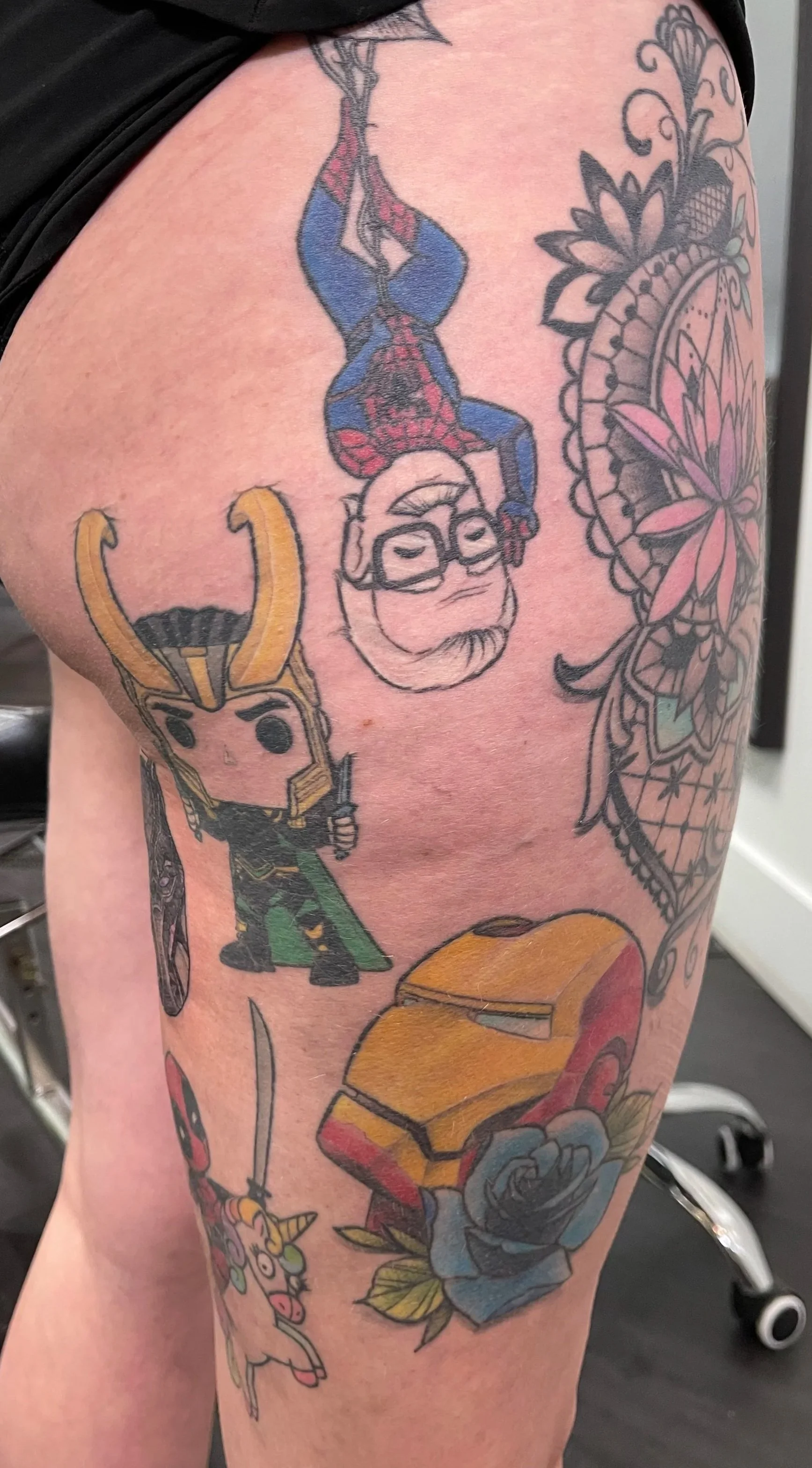 Tattoo on person's thigh featuring a cartoonish Loki with a sword and helmet, a pink and blue unicorn with a rainbow mane, a yellow helmet resembling Iron Man's, and part of a floral and mandala design.