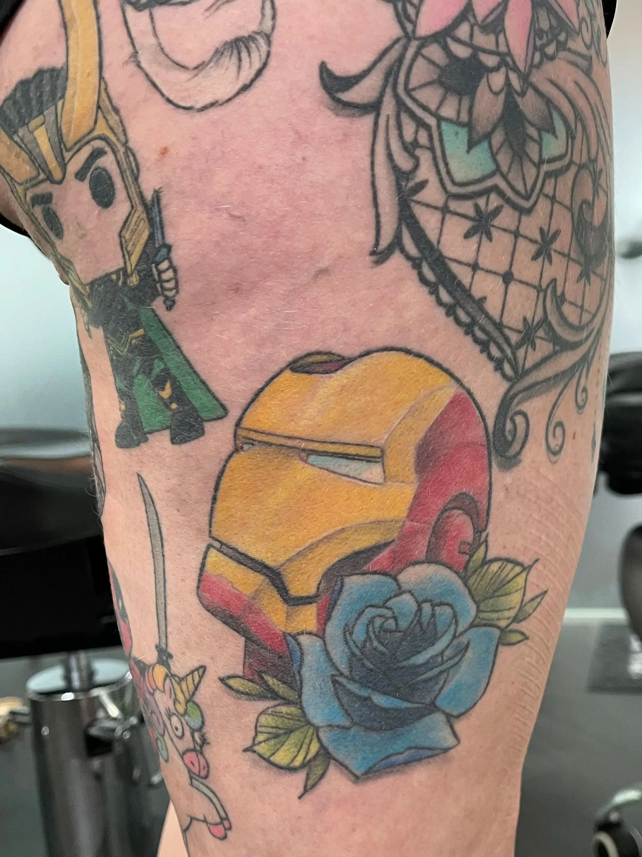 Tattoo of Iron Man helmet with a blue rose below it on a person's upper arm.
