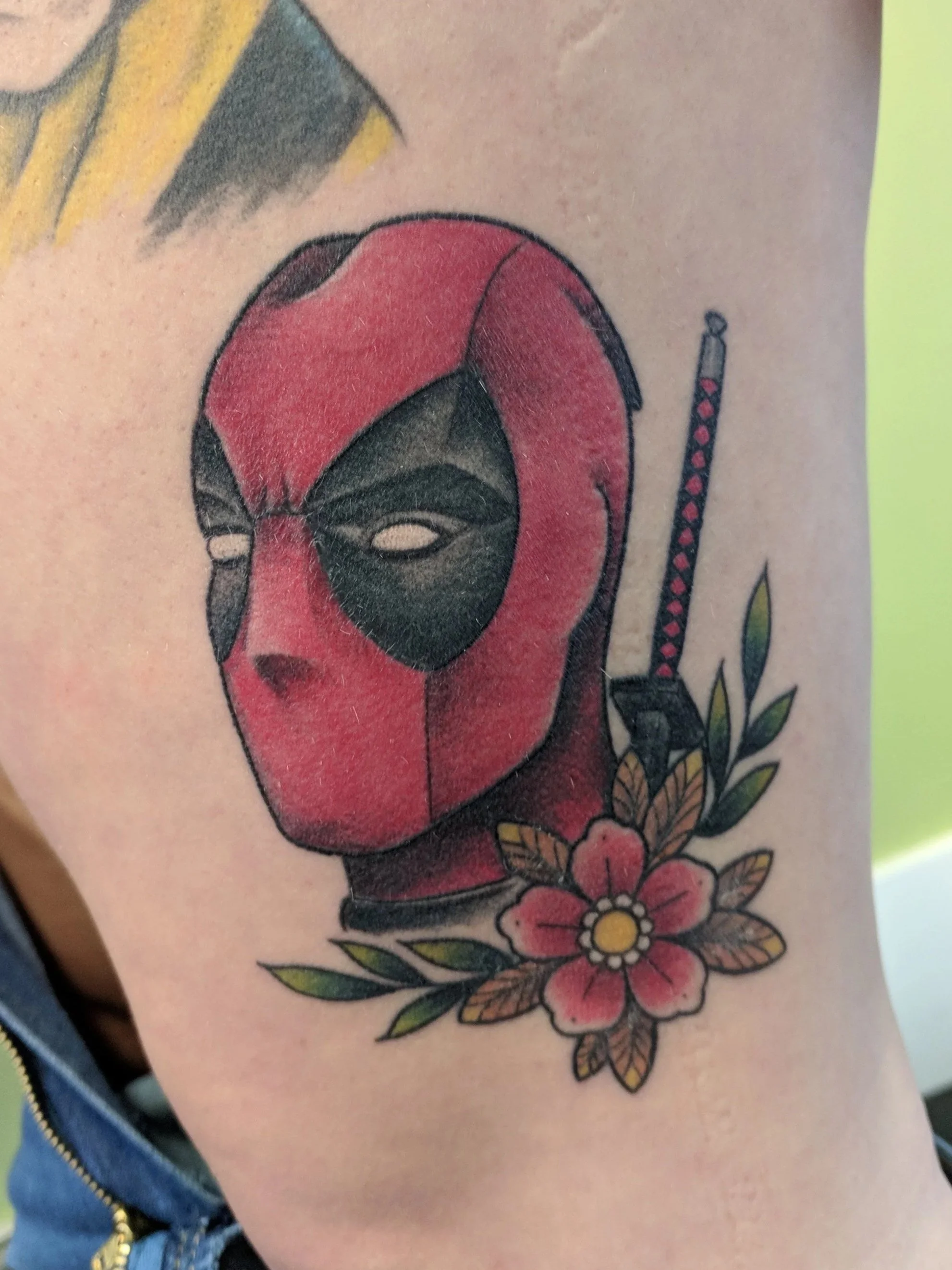 Tattoo of Deadpool's face wearing a red mask with black eye patches, surrounded by a pink flower with green leaves, and a sword with a red and black handle behind it.