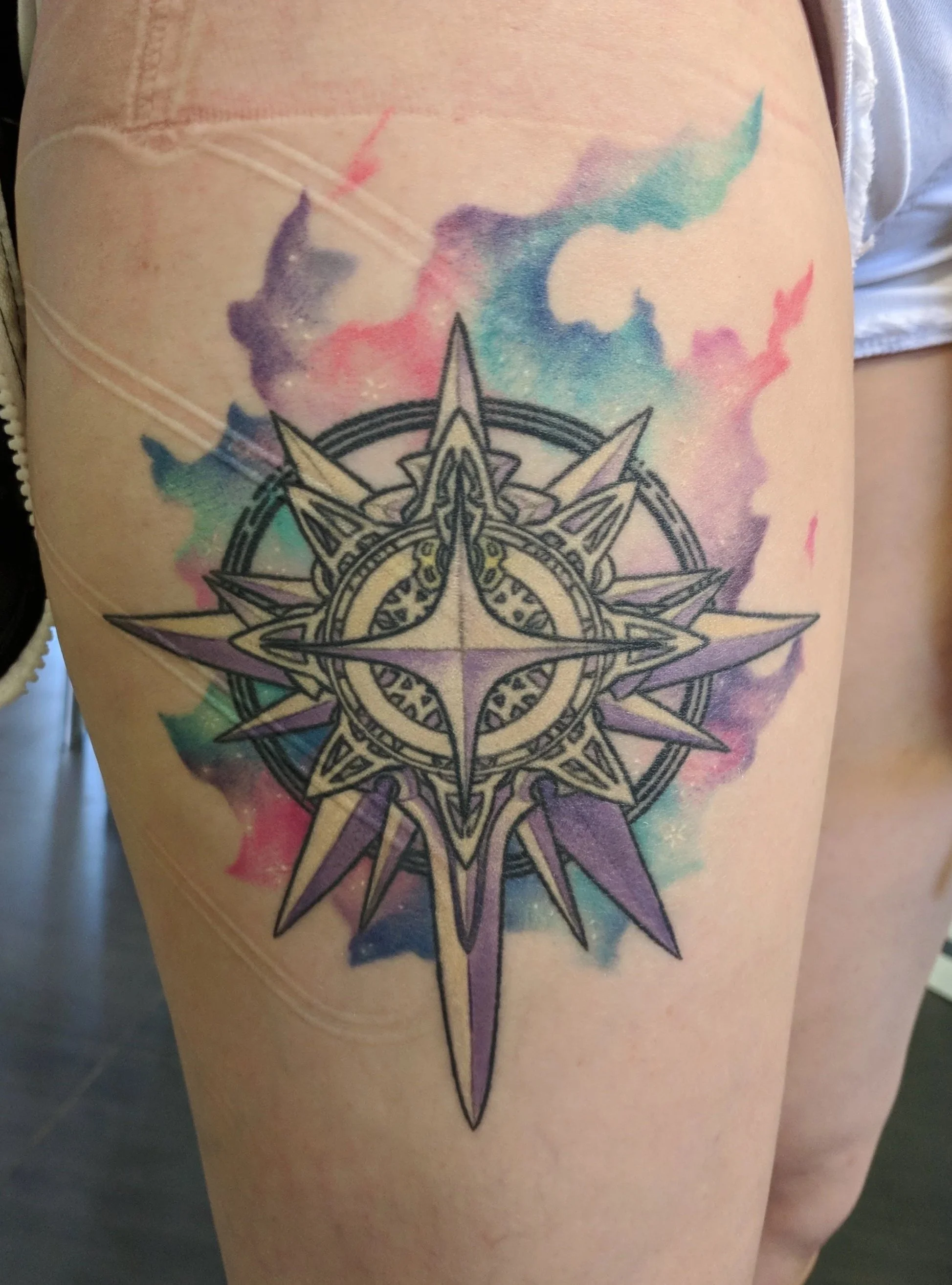 Tattoo of a compass rose with watercolor background on person's thigh.