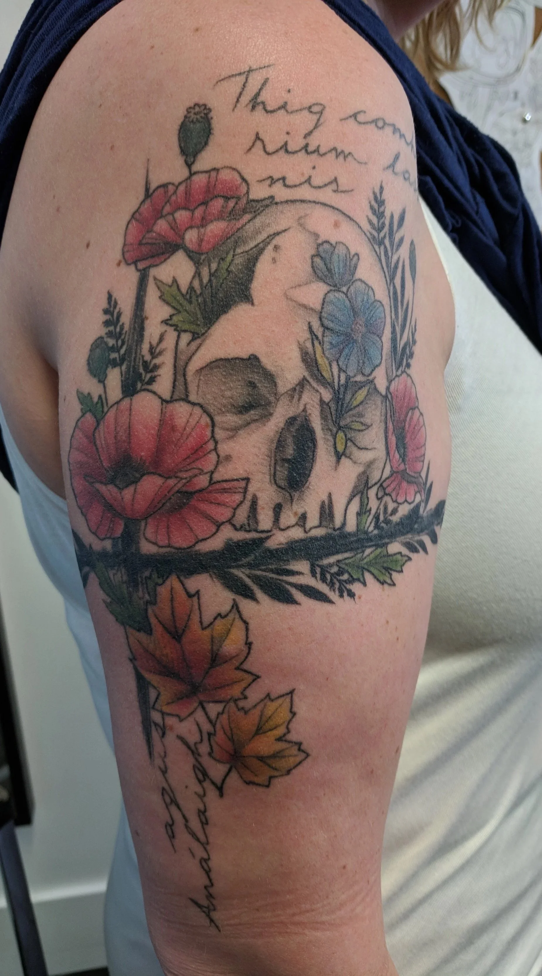 Tattoo of a skull surrounded by colorful flowers and leaves on a person's upper arm, with handwritten text at the top reading 'This comes from he kindest' and cursive script on the lower part reading 'thankful'.