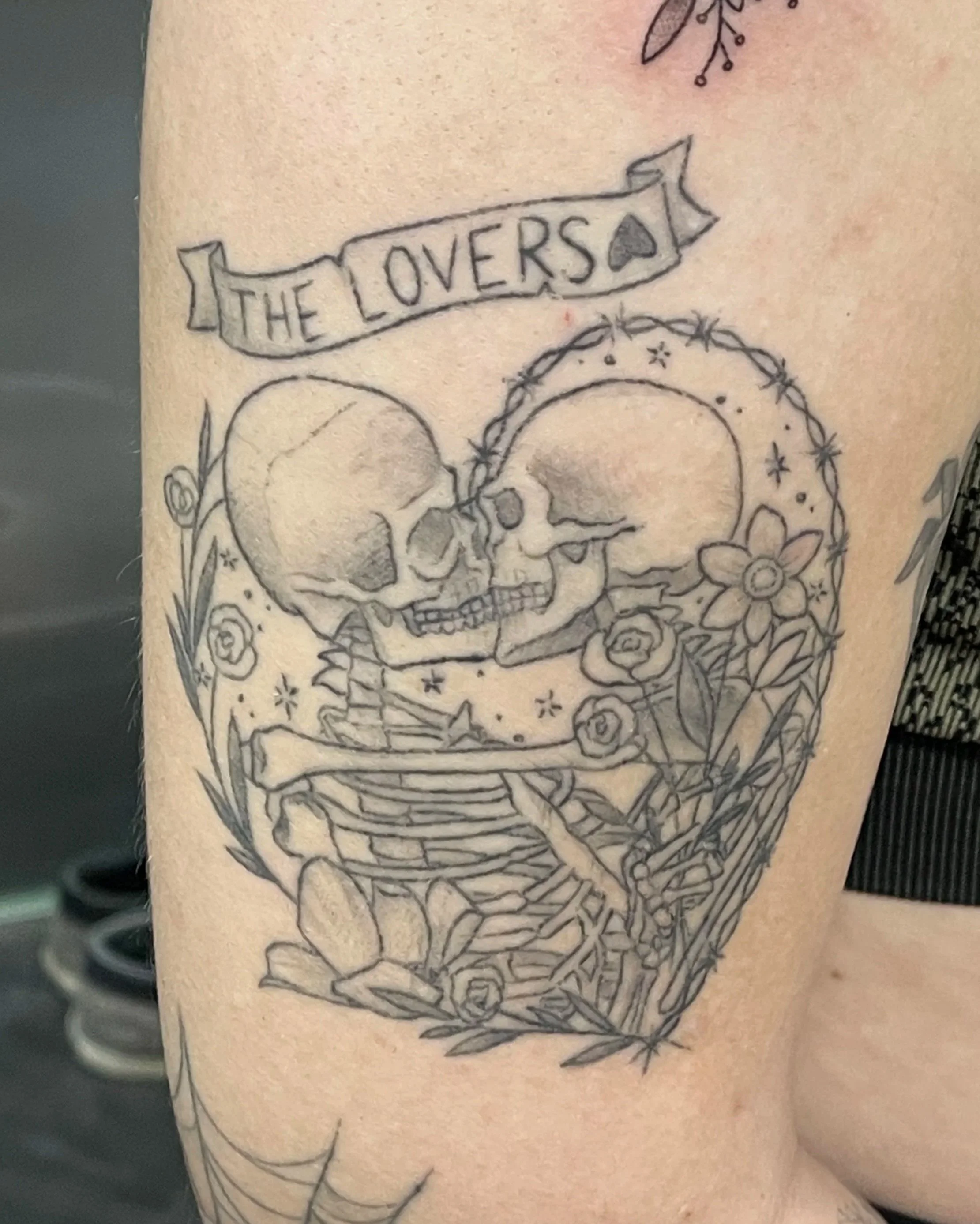 A tattoo of two skulls facing each other, surrounded by flowers and a wreath, with a banner reading 'THE LOVERS' above them, and a spider web at the bottom.
